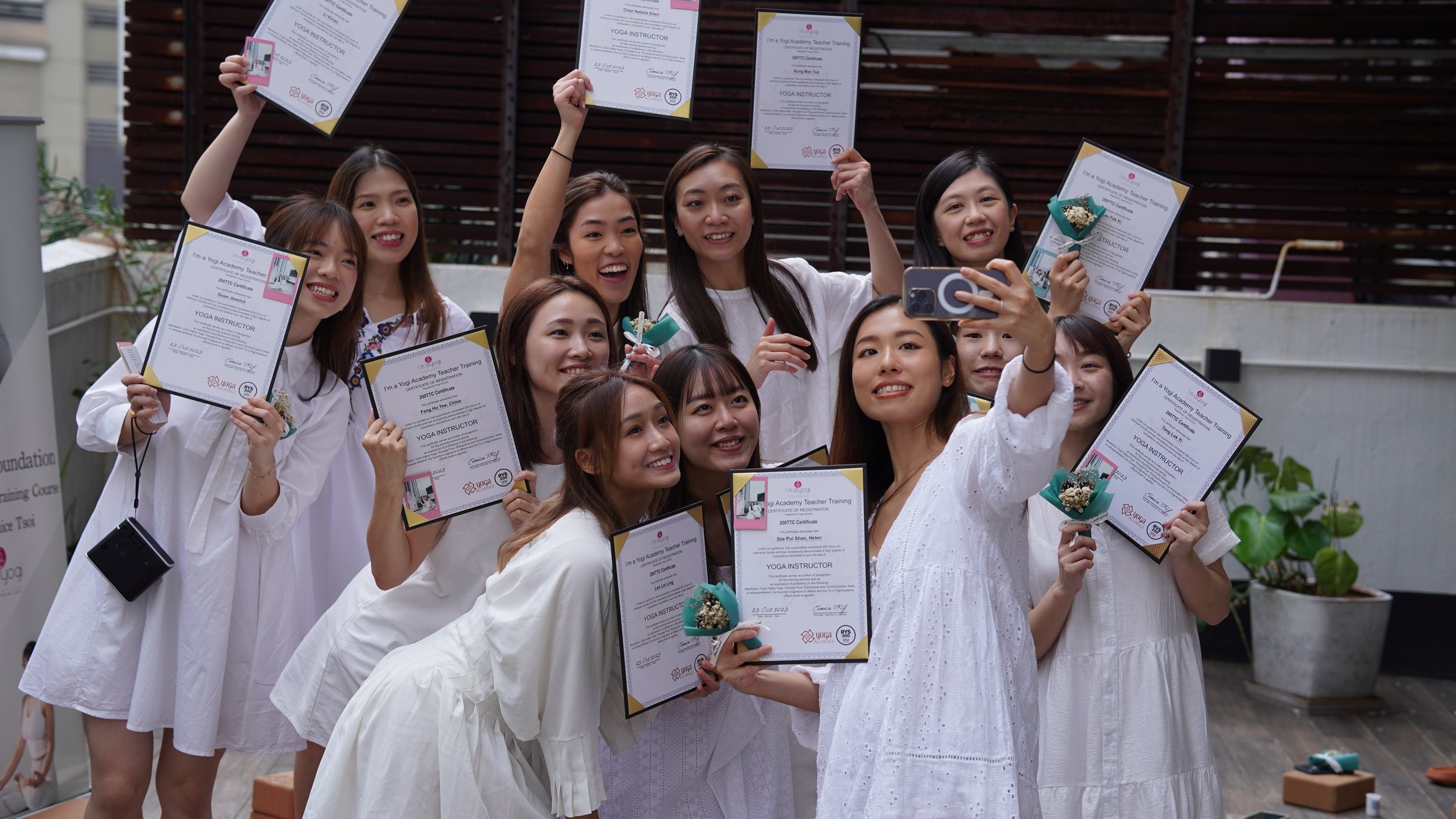 Group of women holding certificates and flowers, taking a selfie together in an outdoor setting.