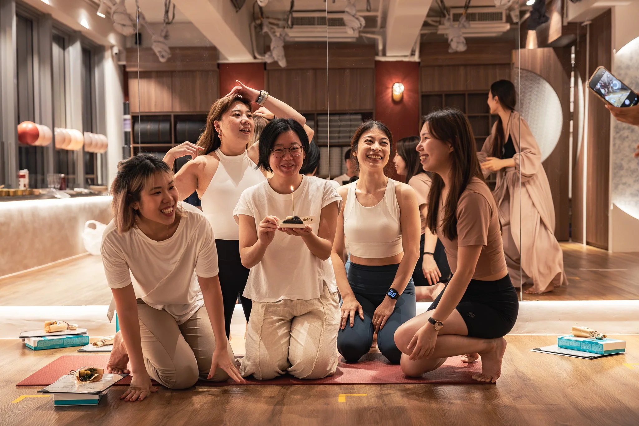 A group of women engaging in a yoga or meditation session in a dance studio, some kneeling on mats, around a birthday cake in the center. All are smiling and appear joyful, with a mirror reflecting the scene and other women in the background.