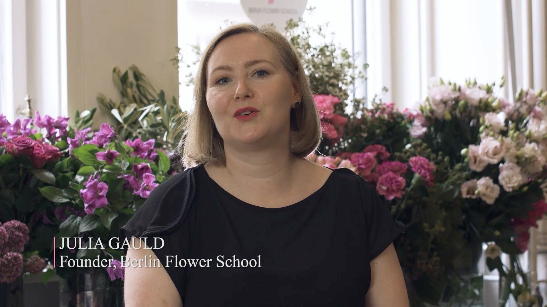 Julia Gauld, founder of Berlin Flower School, speaking in a flower shop with large arrangements of pink and white flowers in the background.