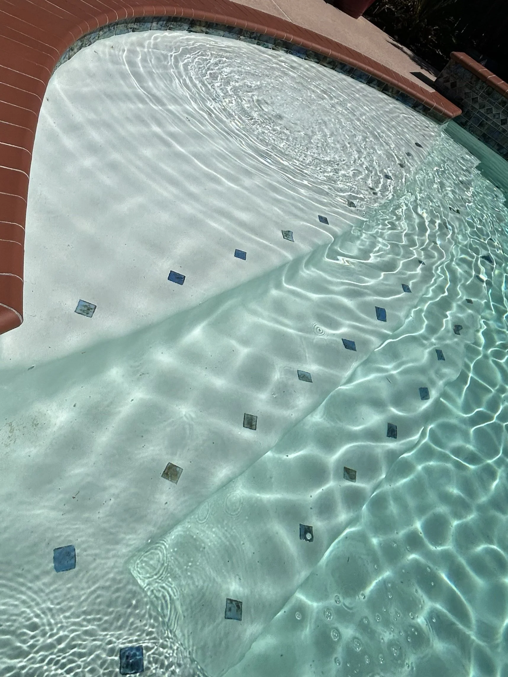 Close-up view of a swimming pool with clear water, small square blue tiles on the bottom, and a red brick edge around the pool.