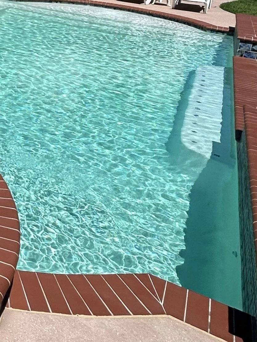An in-ground swimming pool with clear blue water, red brick coping, and white stairs leading into the pool. There are white chairs and lush green grass nearby.
