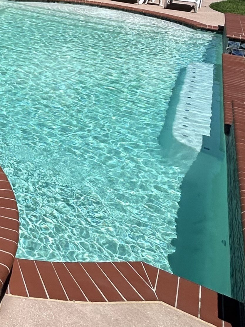 An in-ground swimming pool with clear blue water, red brick coping, and white stairs leading into the pool. There are white chairs and lush green grass nearby.
