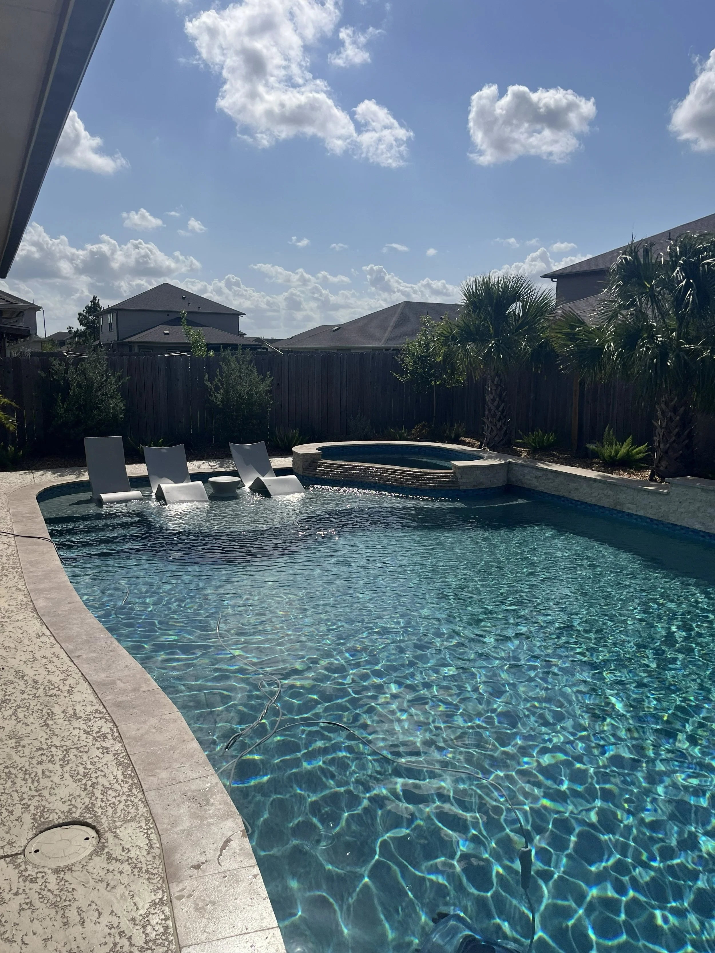 Residential backyard with a swimming pool, pool lounge chairs, palm trees, and neighboring houses under a partly cloudy sky.