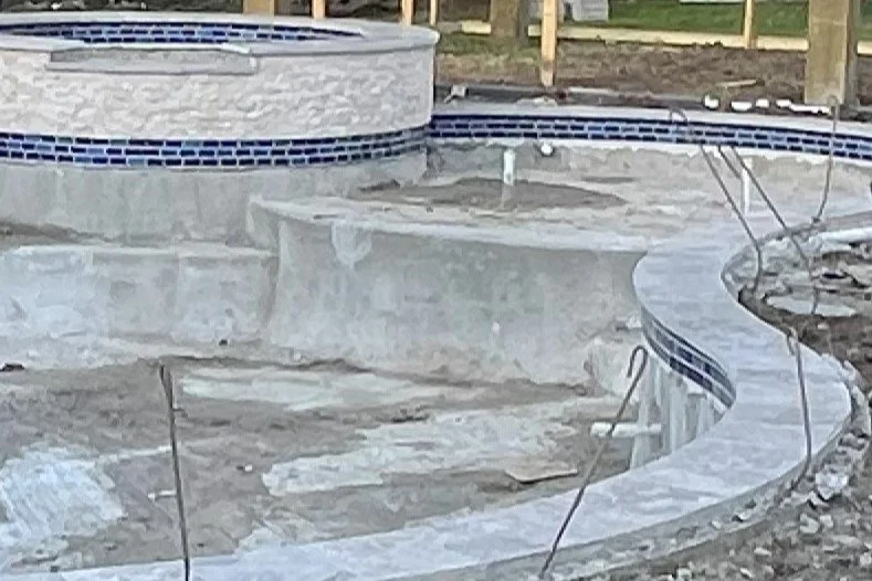 Under construction swimming pool with a curved shape, concrete surface, and partially installed tiles, surrounded by construction wiring.