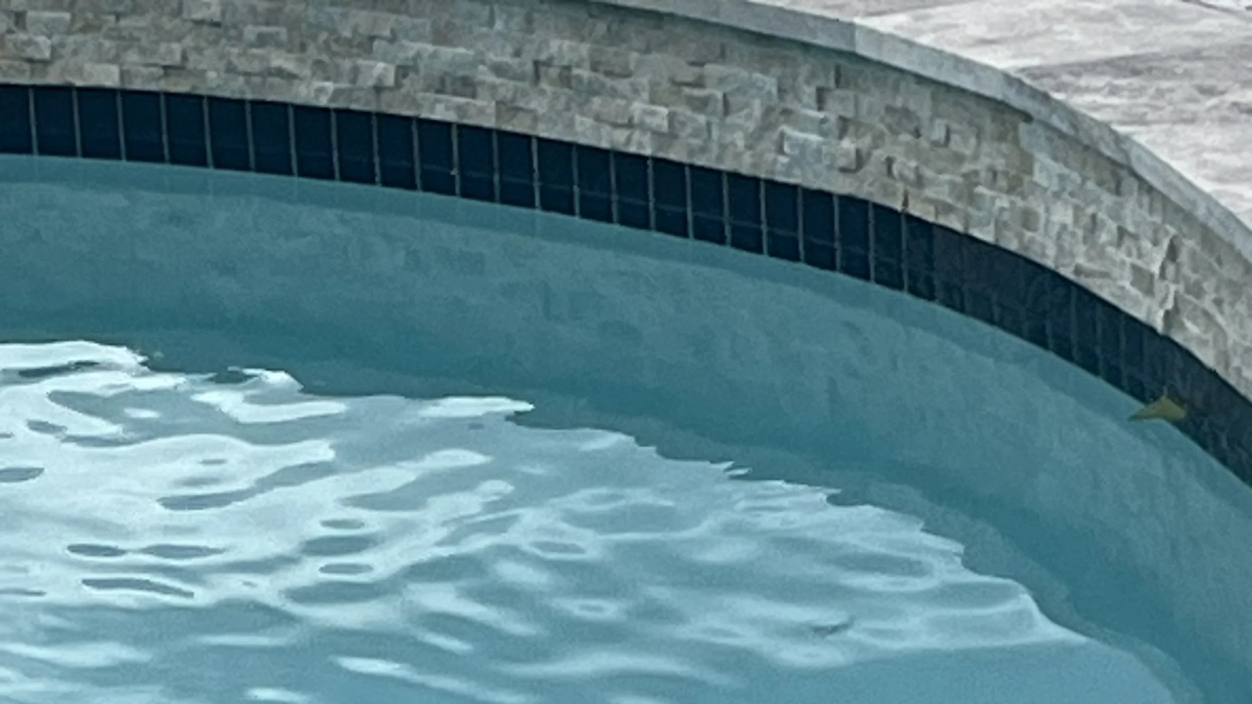 Close-up of a swimming pool featuring new NPT Stonescapes Mini-pebble plaster in White Osprey, new travertine coping, and new NPT Moonscapes waterline tile in Steel Blue, a new spray deck over Carvestone.