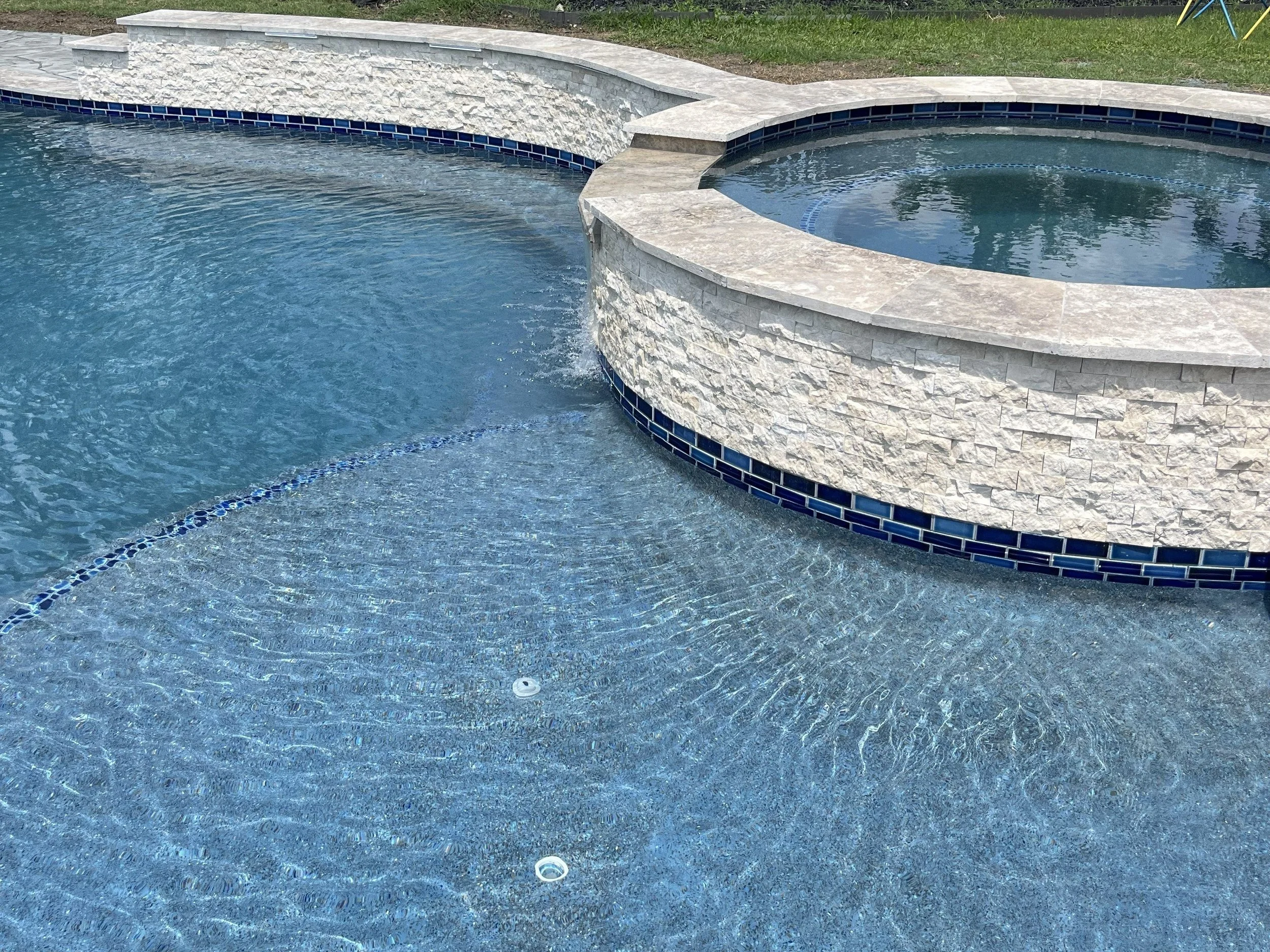 A swimming pool with a raised hot tub, both edged with stone tiles and a decorative blue tile border, and surrounded by grass.