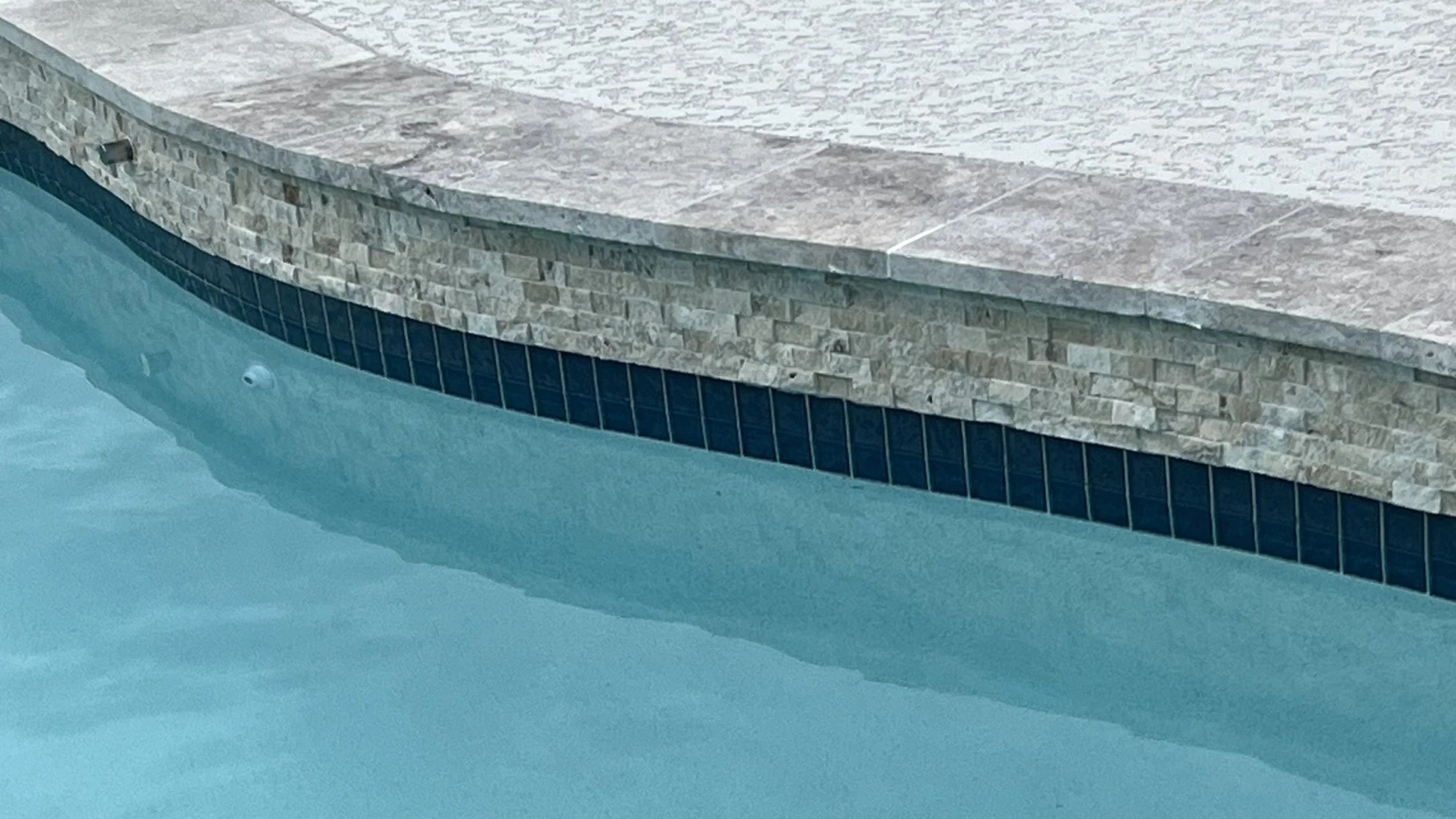 Close-up of a pool featuring new NPT Stonescapes Mini-pebble plaster in White Osprey, new travertine coping, and new NPT Moonscapes waterline tile in Steel Blue, a new spray deck over Carvestone.