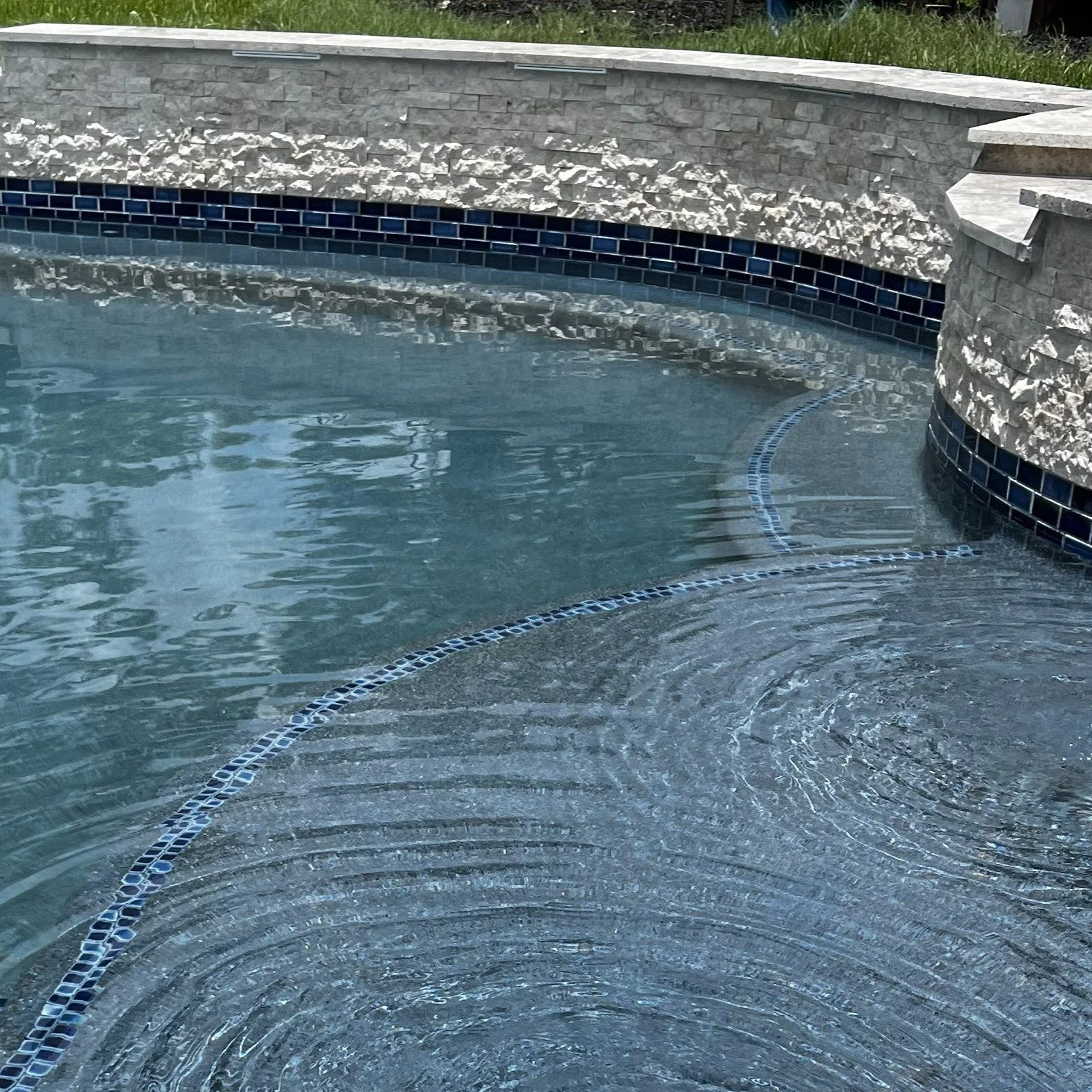 Close-up of a swimming pool with water ripples, tiled edge, and stone wall surround.
