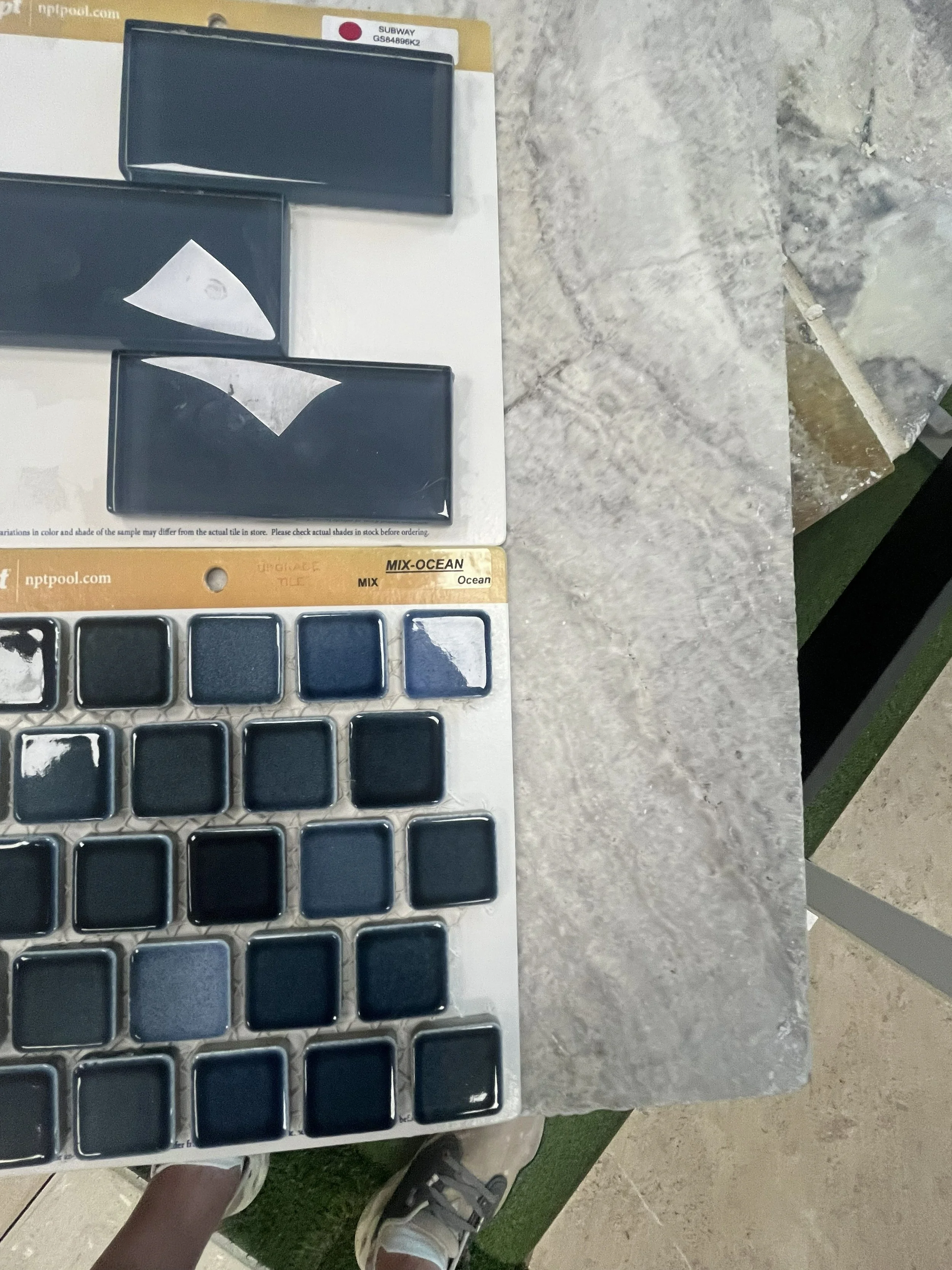 Color swatch sample cards labeled 'MIX-OCEAN' and 'SUBWAY' on a display board on a stone table, with a person's feet visible in sneakers and shorts in the background.