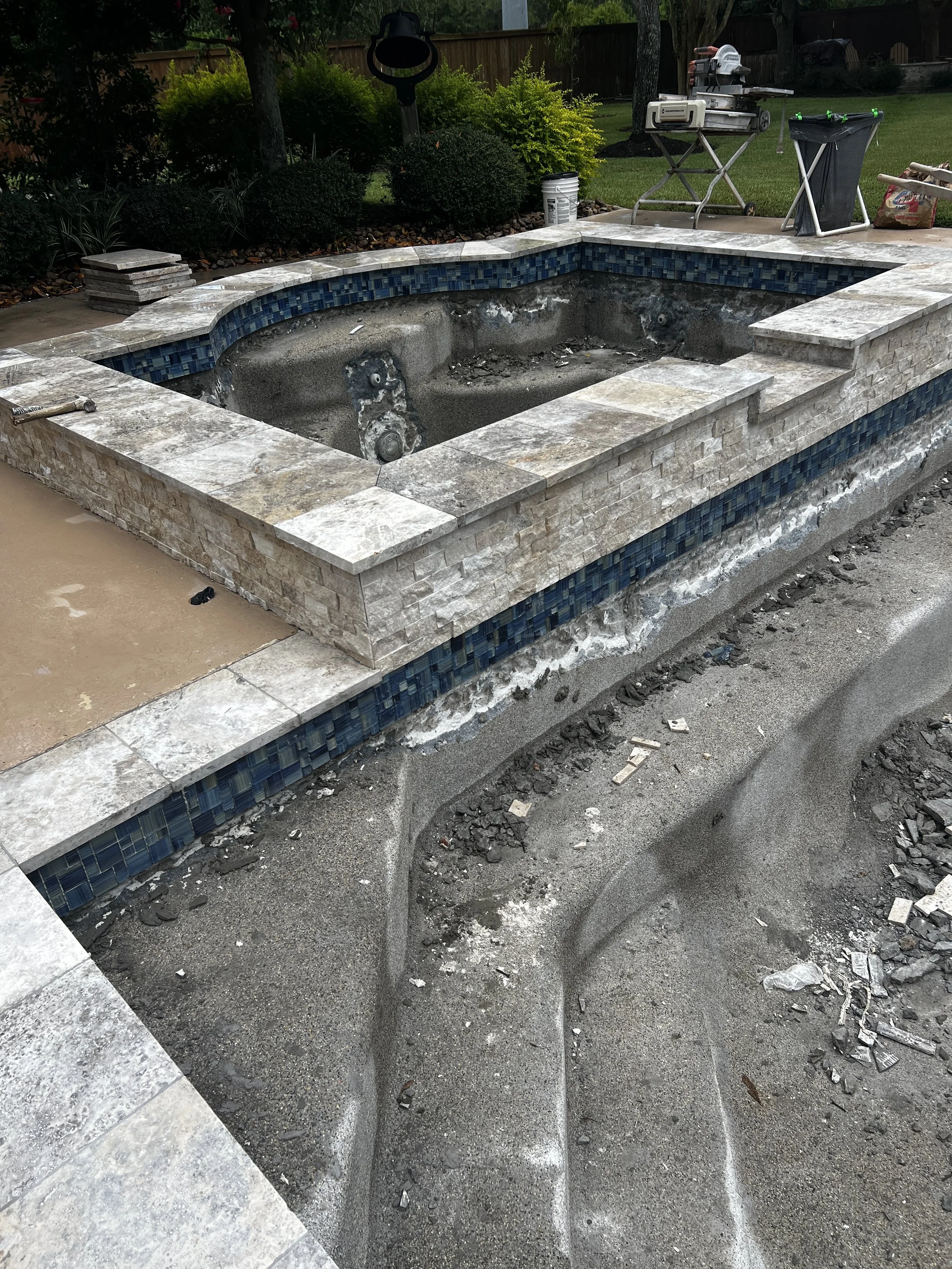 In-ground hot tub under construction with stone edging and blue tile accents, surrounded by dirt and construction debris, backyard with trees and fencing in the background.