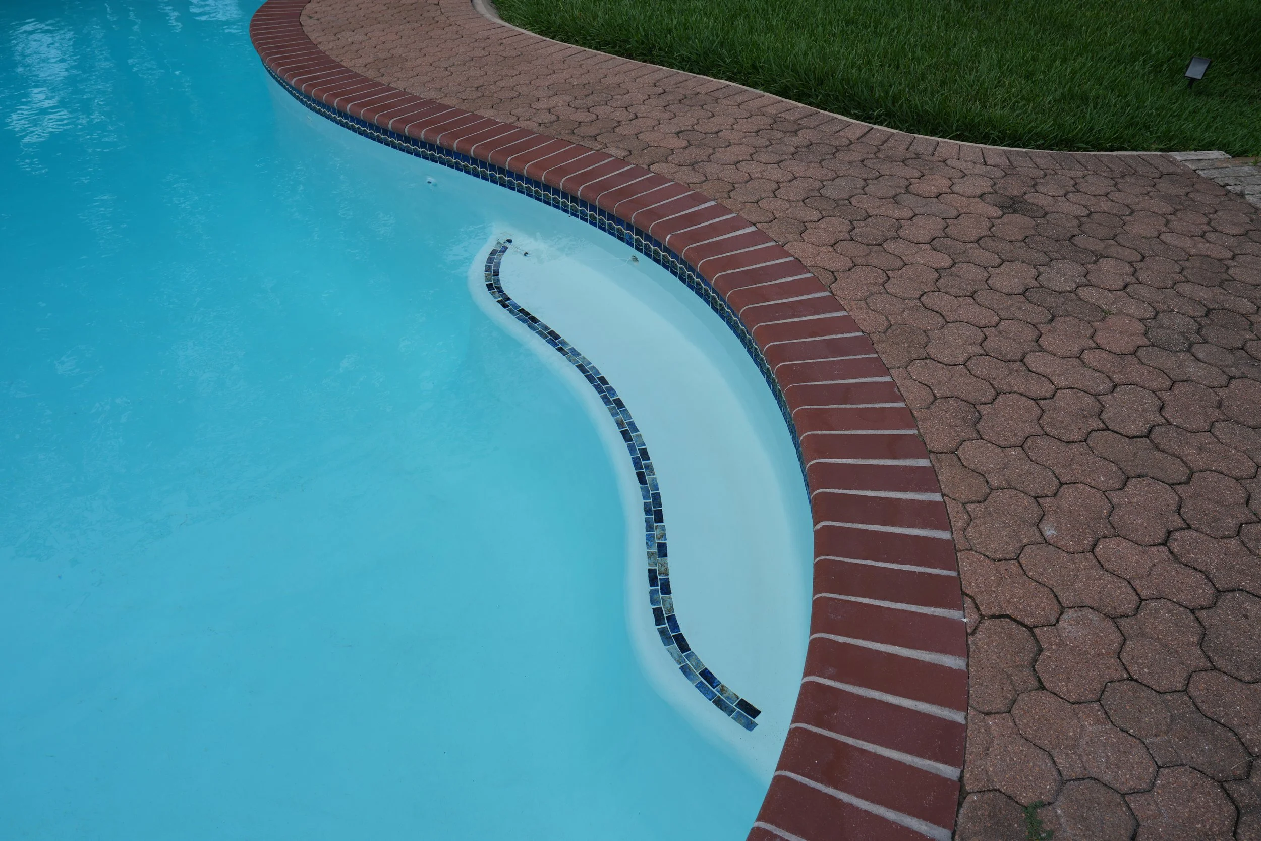 .A picture of a renovated backyard swimming pool surrounded by red brick paving.. Pool features new white plaster with NPT Baroque Blue glossy accent tile and a new pool ladder.