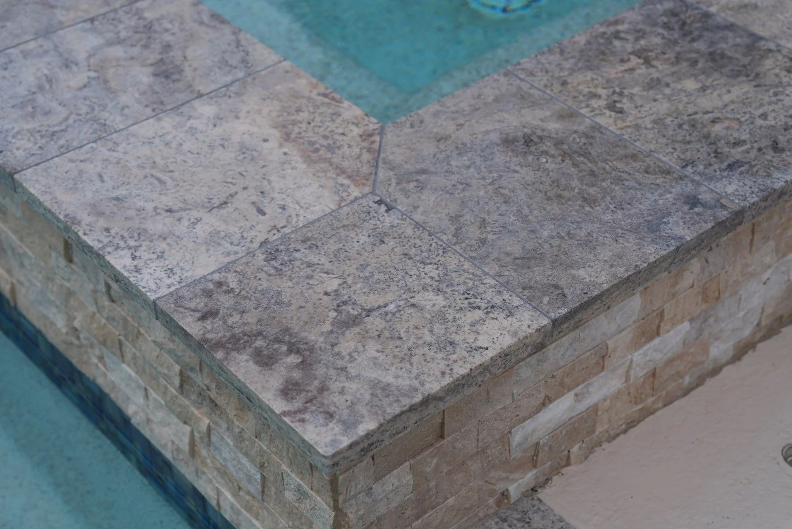 Close-up of silver-toned coping pieces meeting at a spa corner above textured travertine cladding.