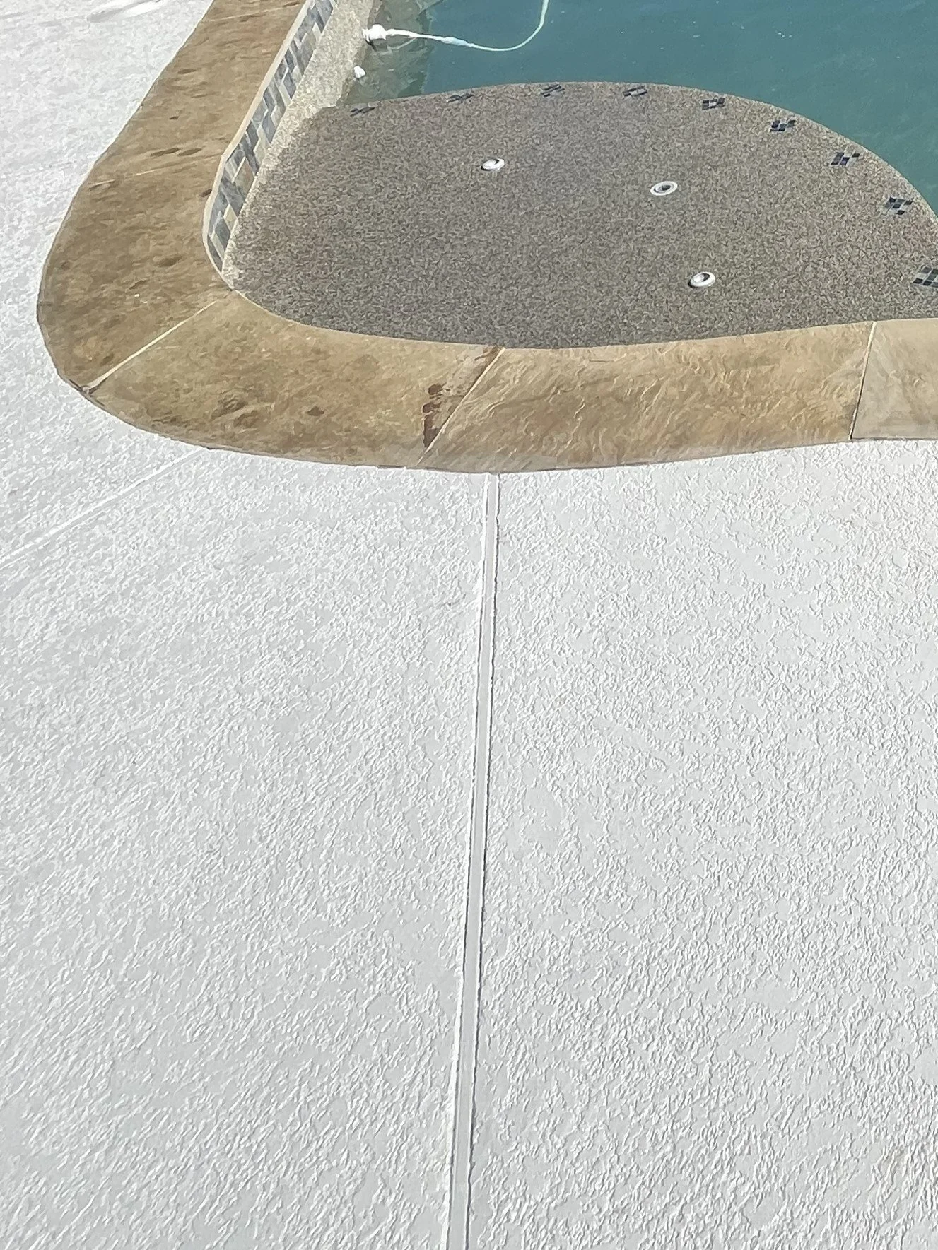 Close-up of a hot tub with a textured gray cover in an outdoor pool area, surrounded by beige tiles and a white textured wall.