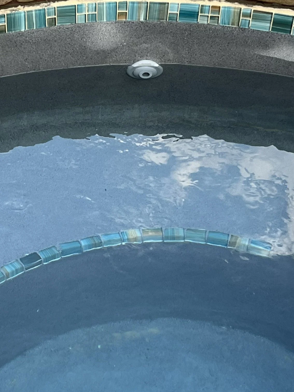 Close-up view of a hot tub with blue mosaic tiles along the edge, water inside reflecting the sky and clouds, and a white light fixture on the wall.
