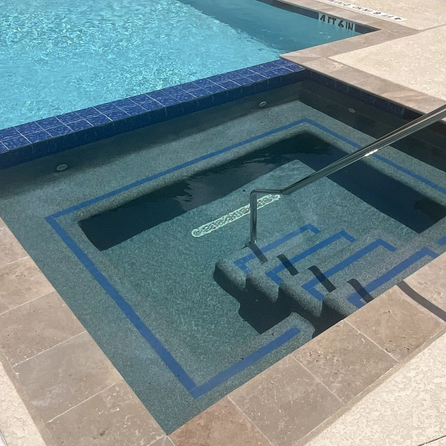 A swimming pool with a hot tub in the foreground, tiled with blue and gray tiles, with steps leading into the hot tub.