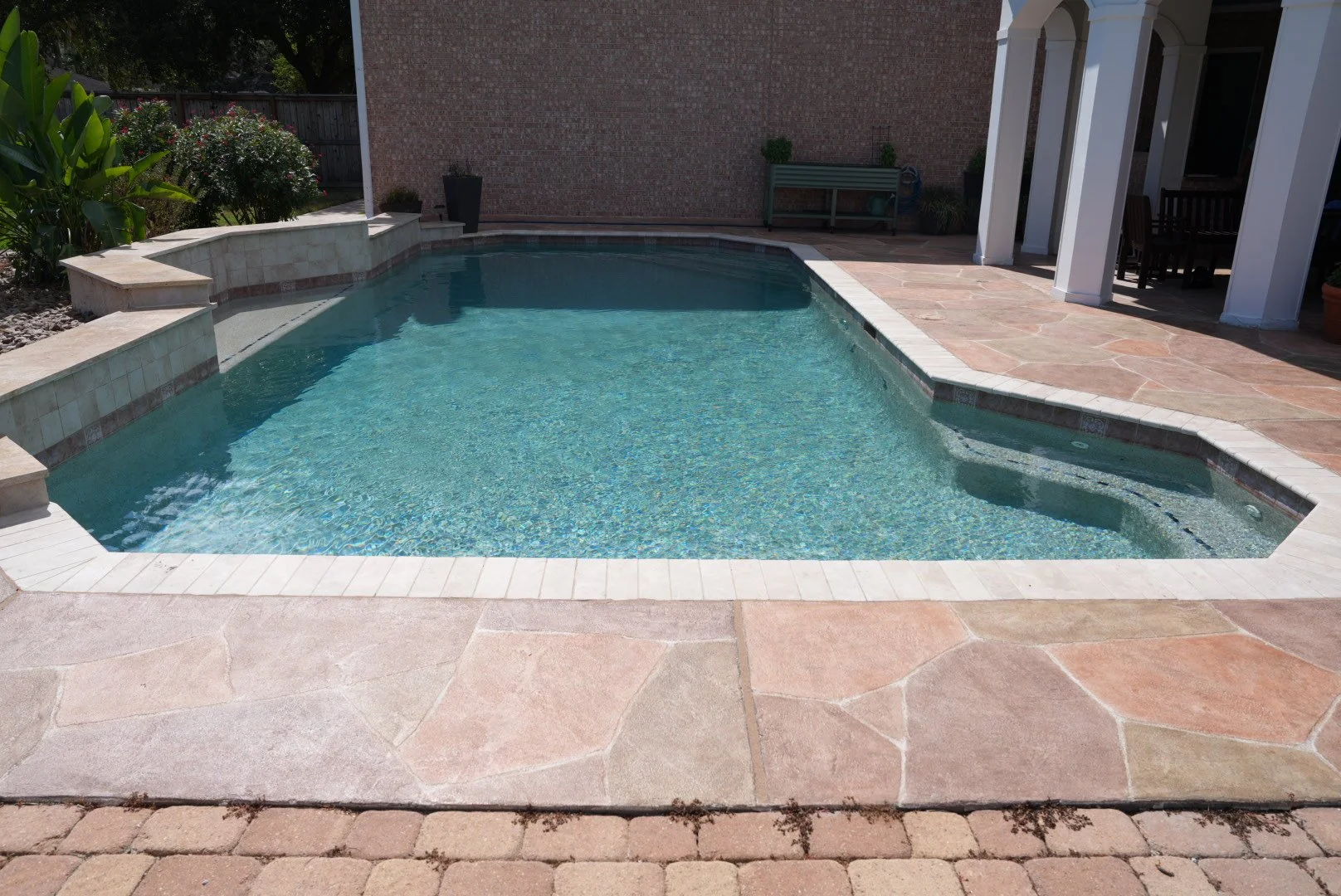 Backyard swimming pool with beige tile lining and tan stone deck, with an adjacent covered patio area with chairs and tables, and a brick wall in the background.