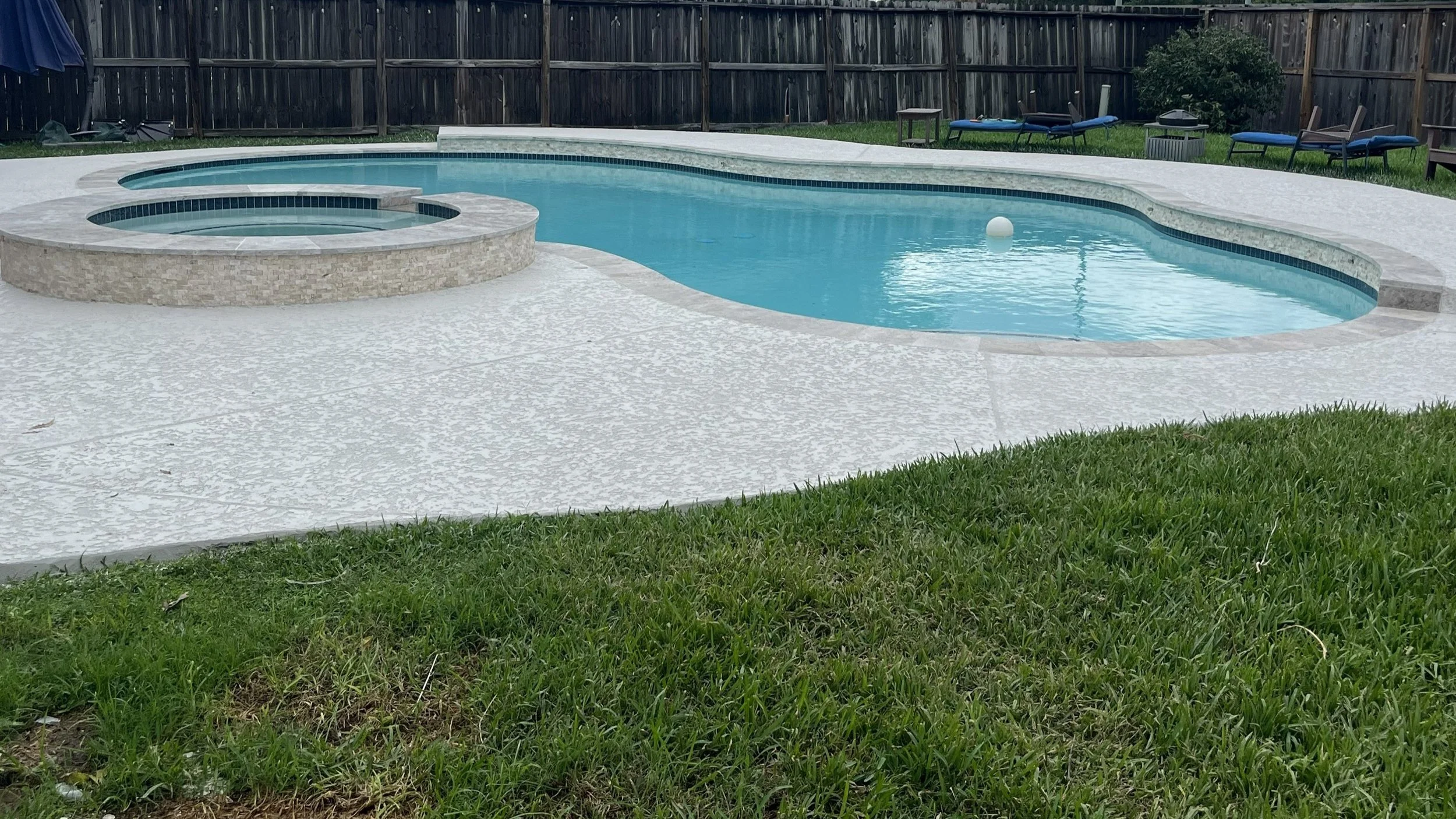 A renovated backyard swimming pool with a spa featuring new NPT Stonescapes Mini-pebble plaster in White Osprey, new travertine coping, and new NPT Moonscapes waterline tile in Steel Blue, a new spray deck over Carvestone.