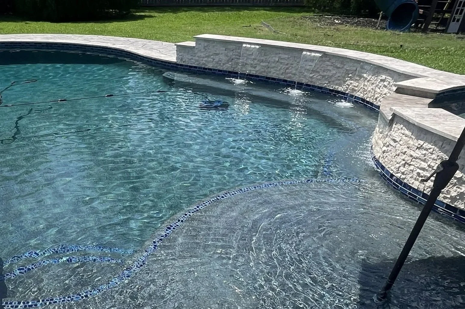 Swimming pool with clear water, stone border, water feature with water cascading from a wall, and a grassy yard in the background.