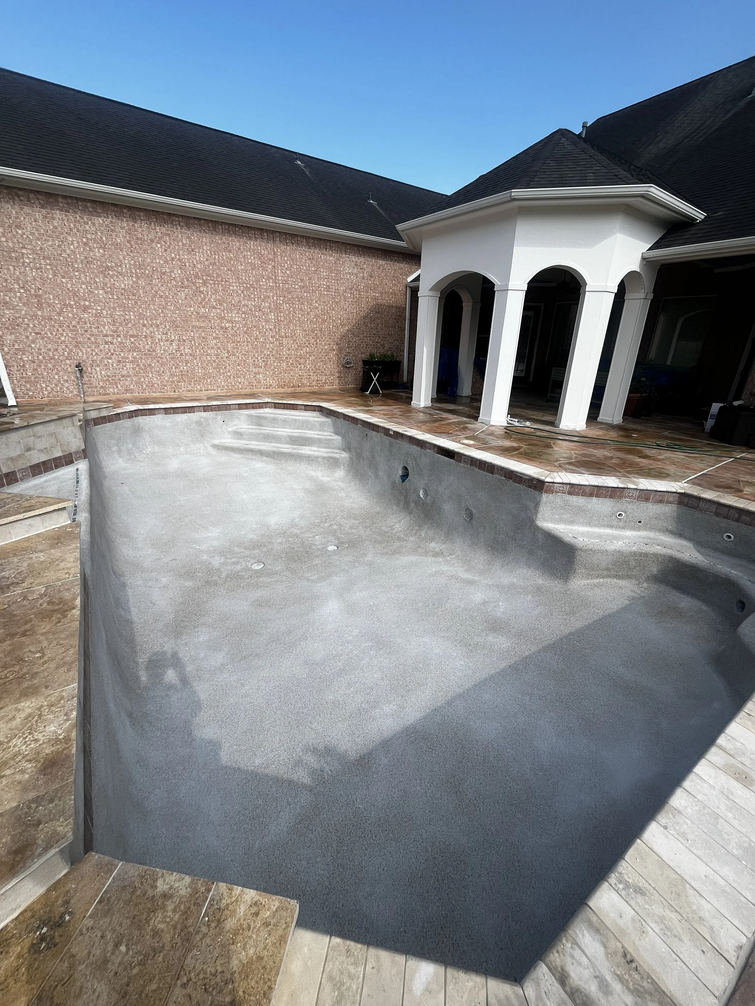 Backyard with empty swimming pool, brick and stone patio, white house with porch, blue sky.