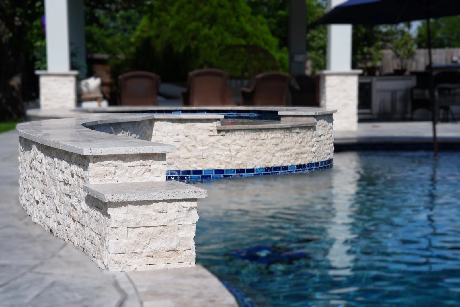 Luxury backyard pool with built-in stone hot tub and blue tile accents, surrounded by a patio and outdoor seating area under a covered patio with umbrellas.