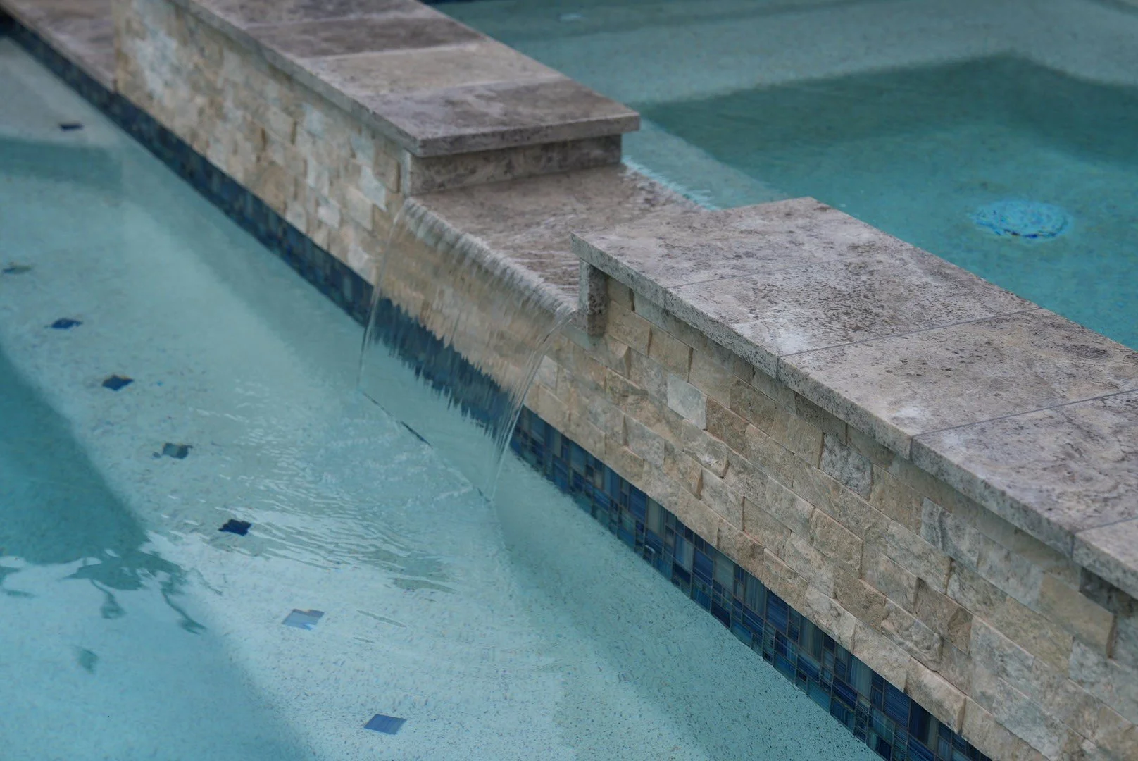 Water flowing over the raised spa spillway above split-face stone cladding and blue mosaic waterline tile.