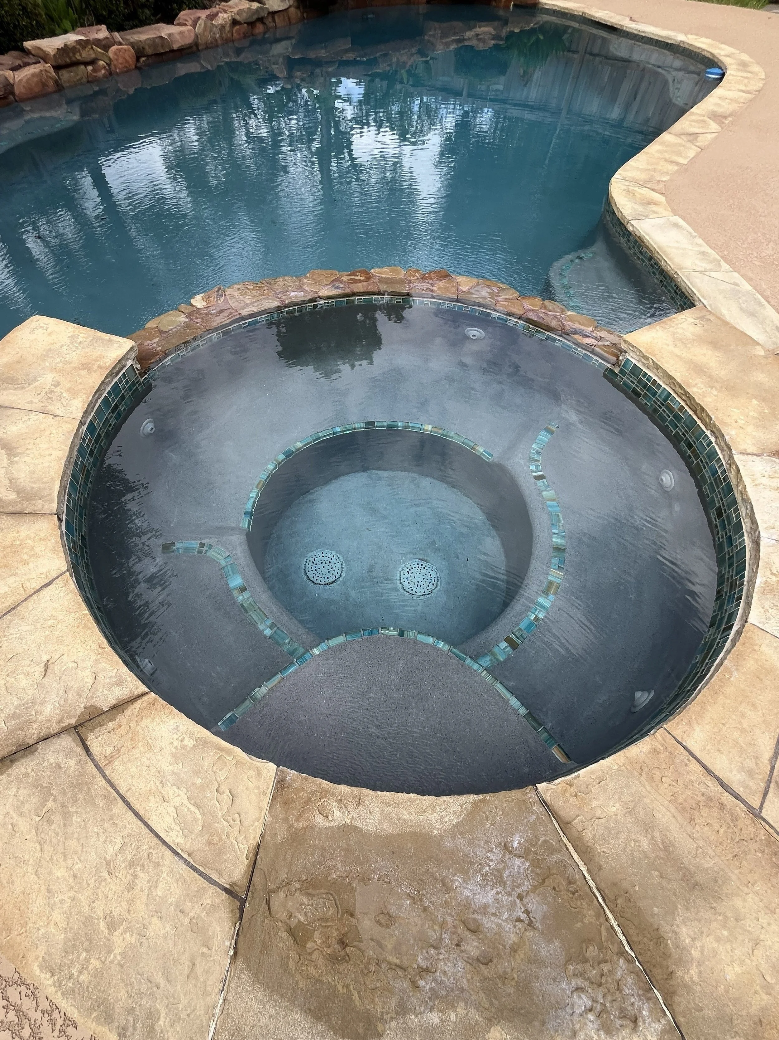 Close-up of a built-in hot tub with water, surrounded by stone paving, overlooking a larger swimming pool with a natural stone border.