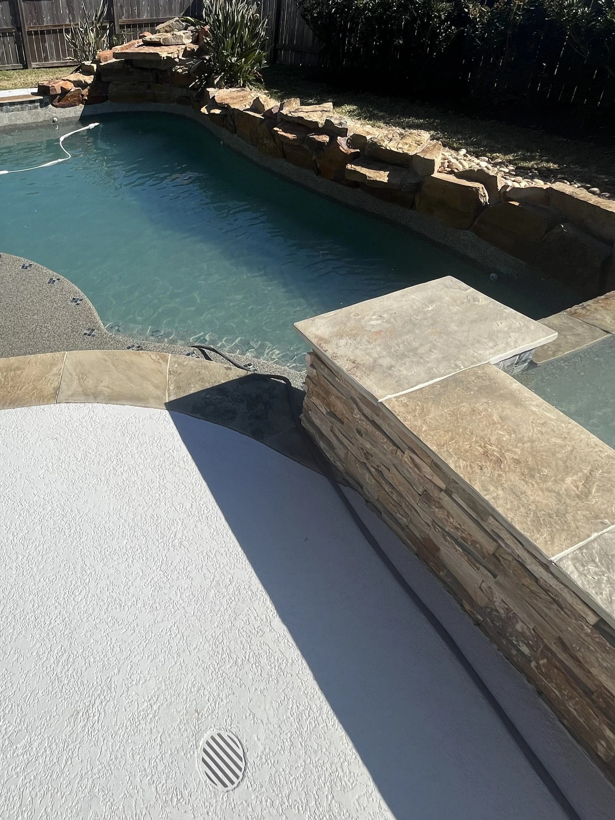 Swimming pool with a rock waterfall feature, surrounded by a stone deck and fencing, under bright sunlight.