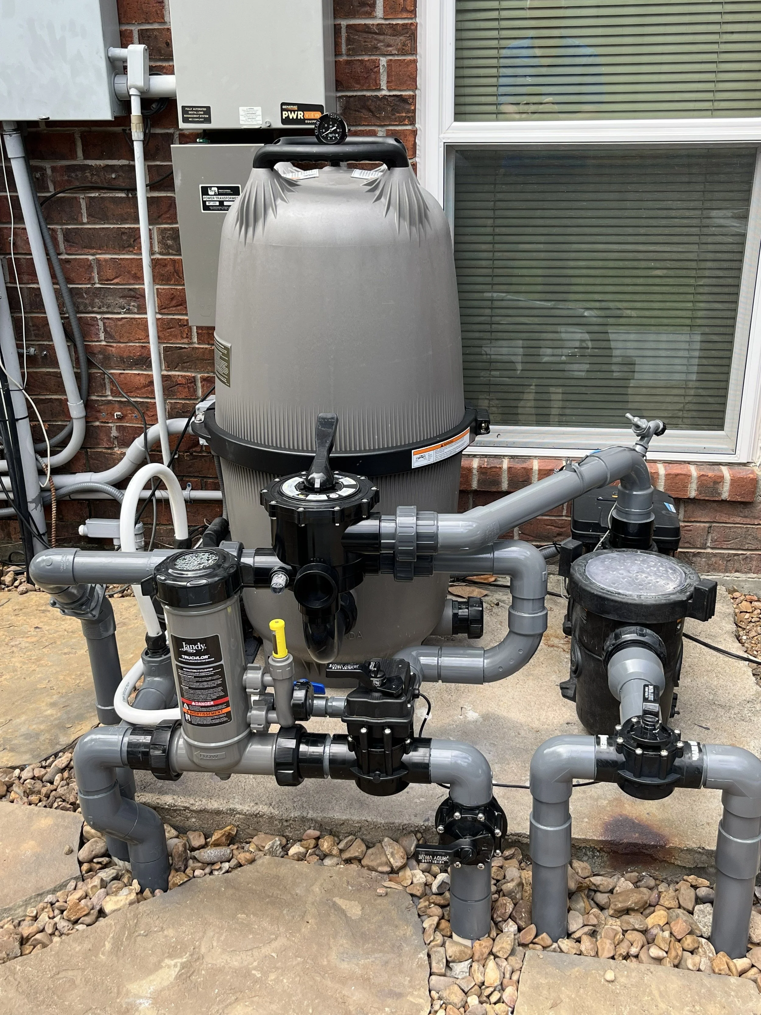 An outdoor pool pump system with filters, control valves, and plumbing pipes installed on a concrete slab near a brick house wall with a window.