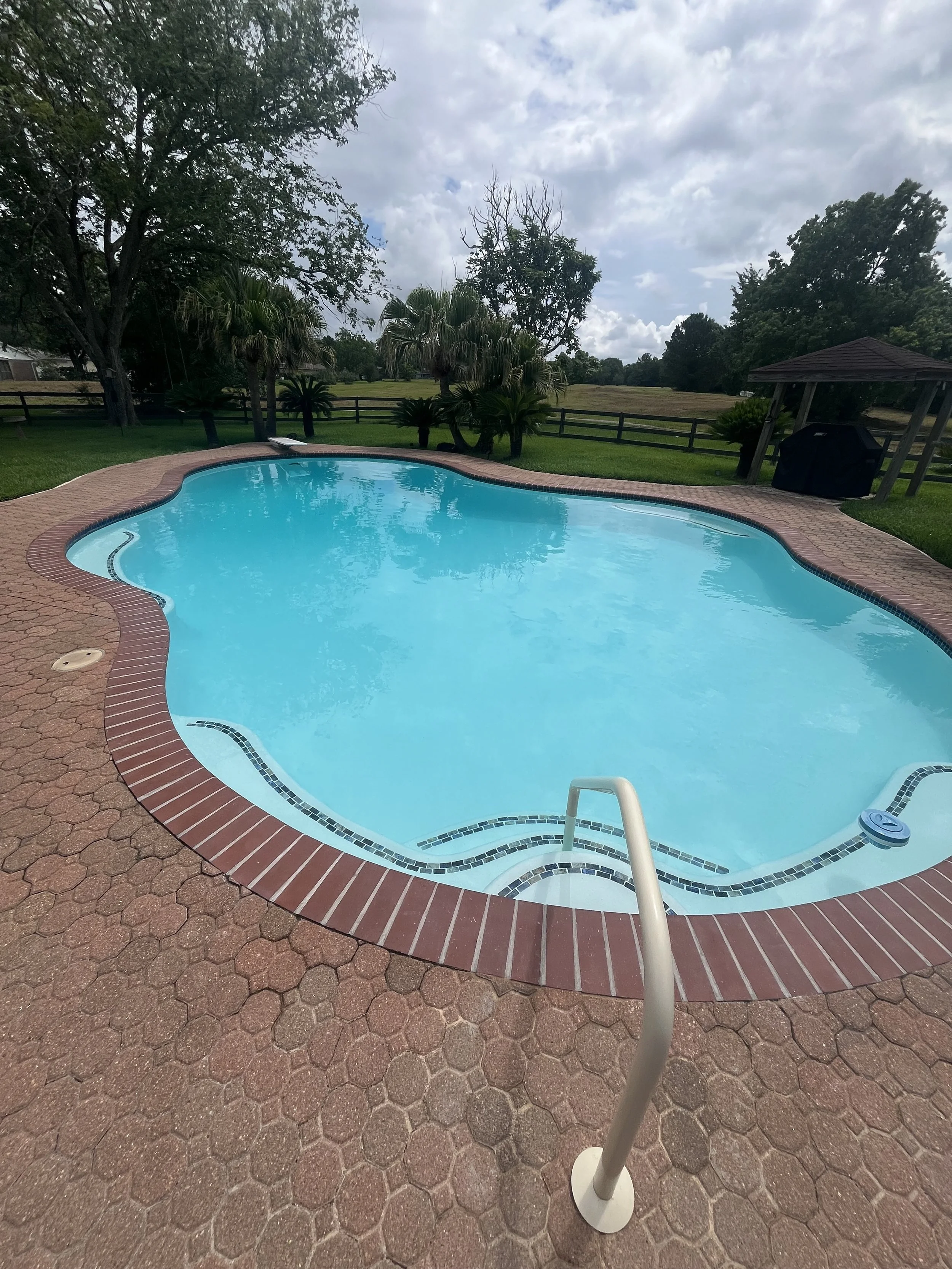 A picture of a renovated backyard swimming pool surrounded by red brick paving and green grass. Pool features new white plaster and a new pool ladder.