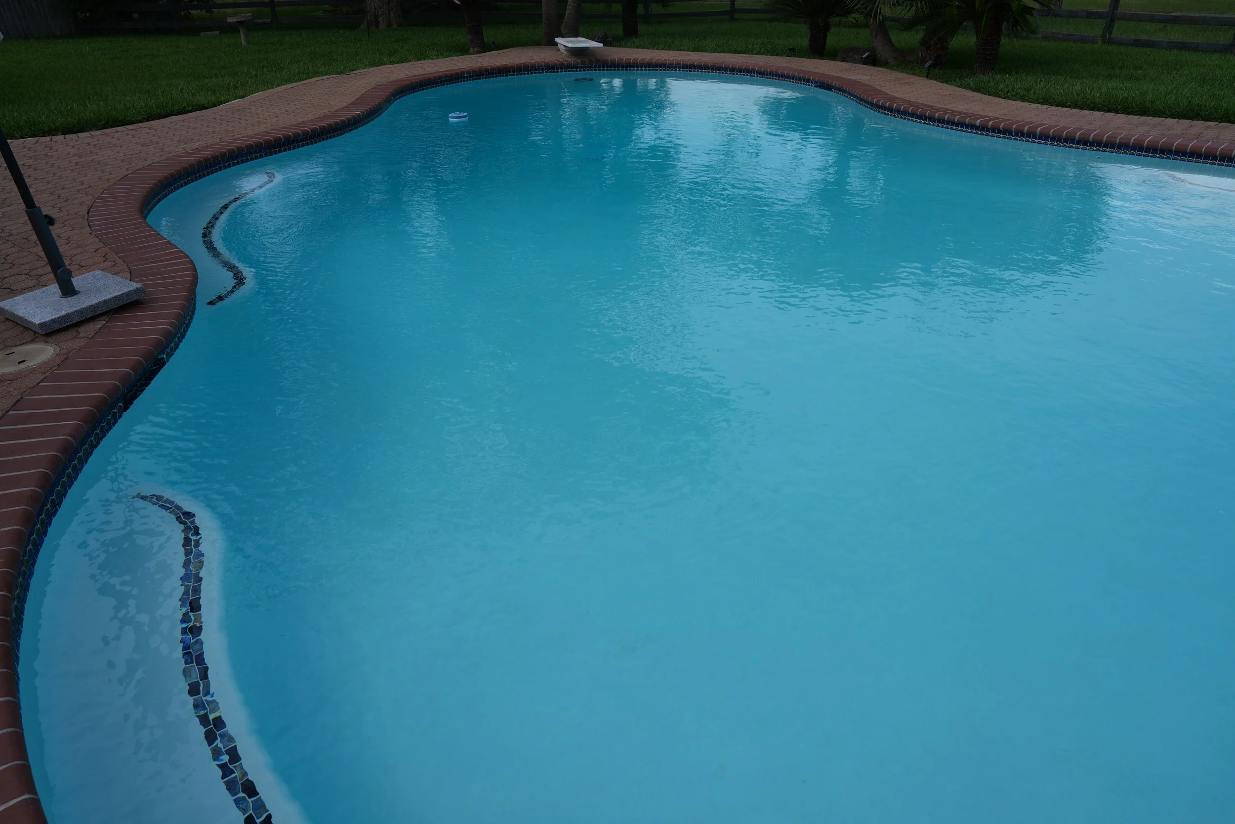 .A picture of a renovated backyard swimming pool surrounded by red brick paving.. Pool features new white plaster with NPT Baroque Blue glossy accent tile and a new pool ladder.