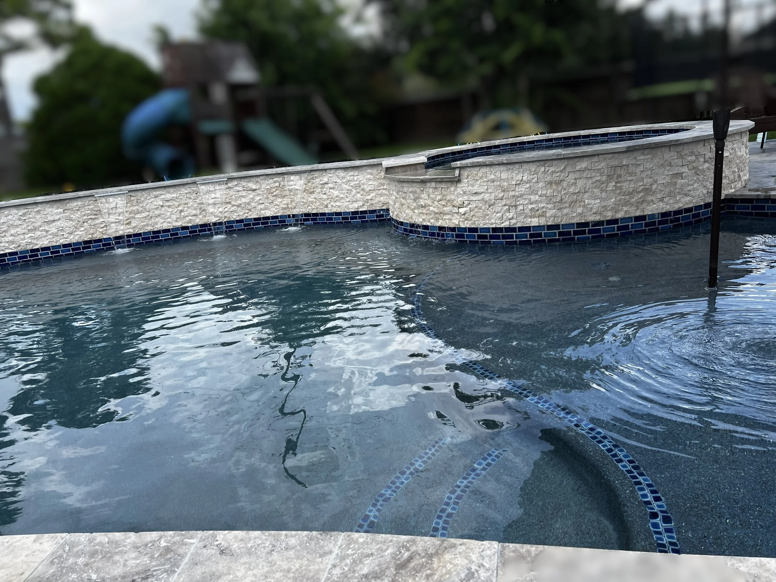 A close-up shot of a hot tub with blue mosaic tiles along the waterline, with a tree and a blue slide in the blurred background.