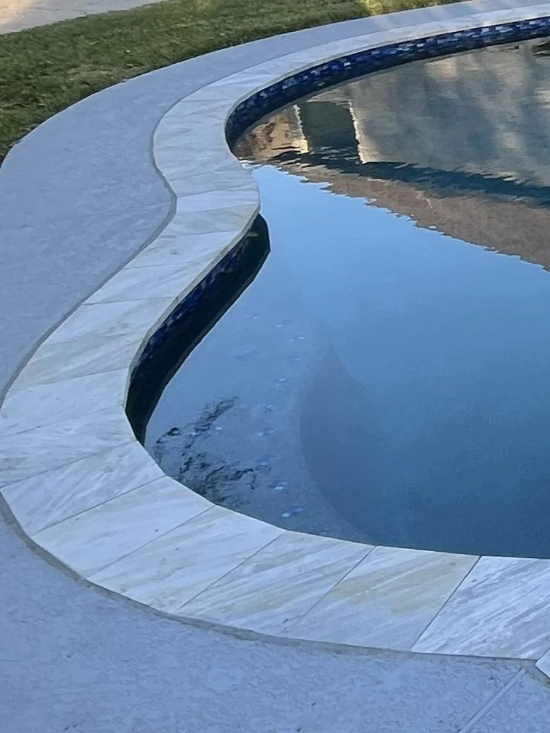 Close-up of a swimming pool with a curved light-colored stone border, reflecting the sky and surroundings in the water.