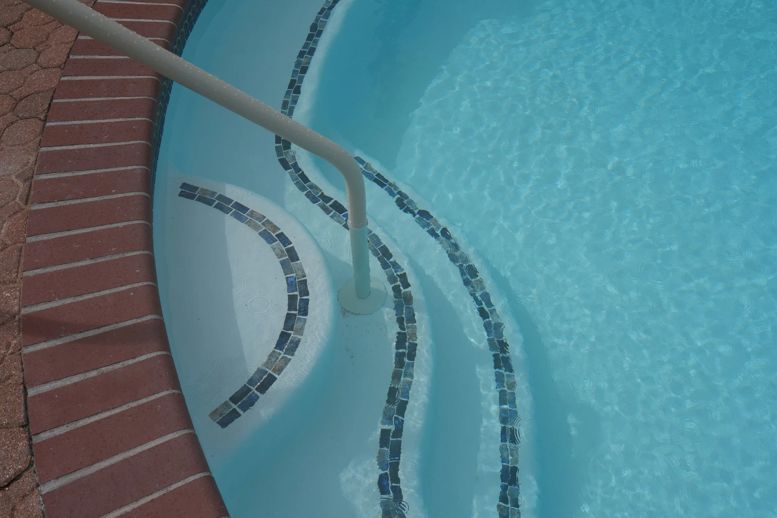.A picture of a renovated backyard swimming pool surrounded by red brick paving.. Pool features new white plaster with NPT Baroque Blue glossy accent tile and a new pool ladder.