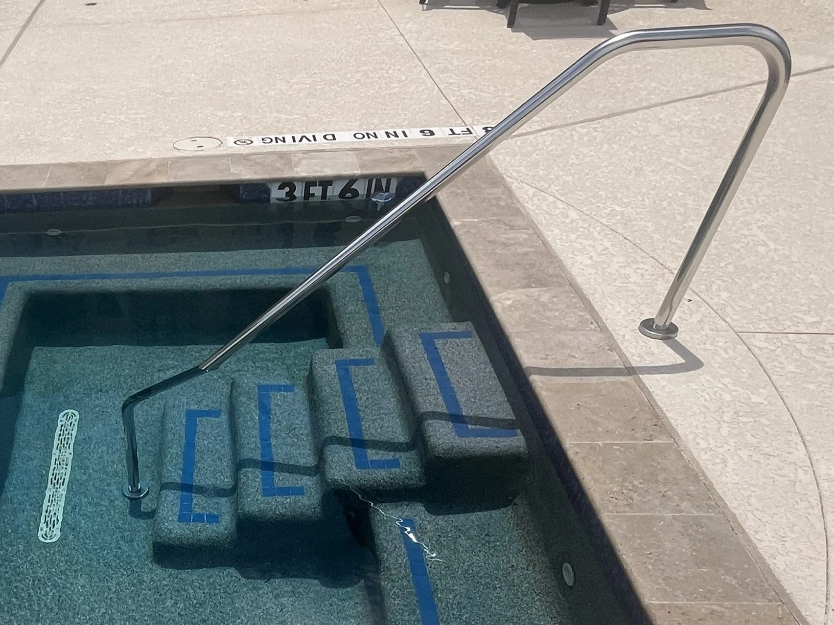 A close-up view of a swimming pool's edge with a metal handrail and a sign indicating a no diving zone. The pool has blue lane markings and floating lane dividers. The pool is surrounded by a tiled deck.