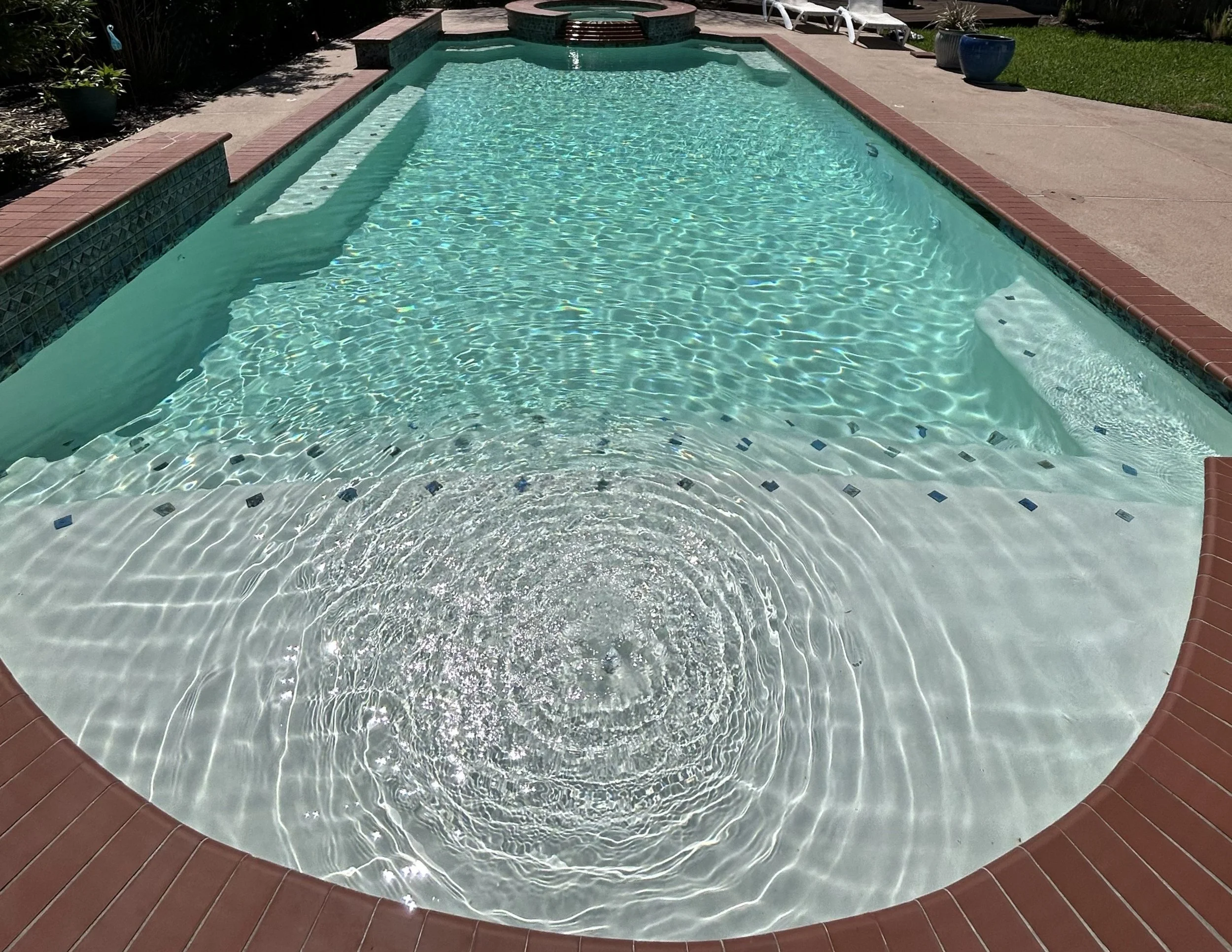 In-ground backyard swimming pool with clear water, surrounded by a brick border and concrete patio, with pool steps visible at one end and lawn with potted plants and lounge chairs in the background.