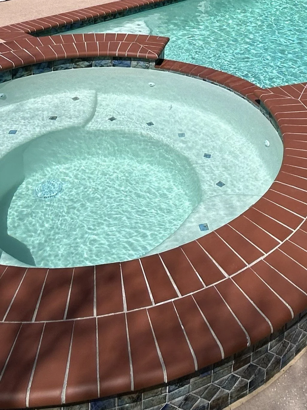 Partial view of a hot tub with clear water, surrounded by a red brick edge, next to a swimming pool with blue water.