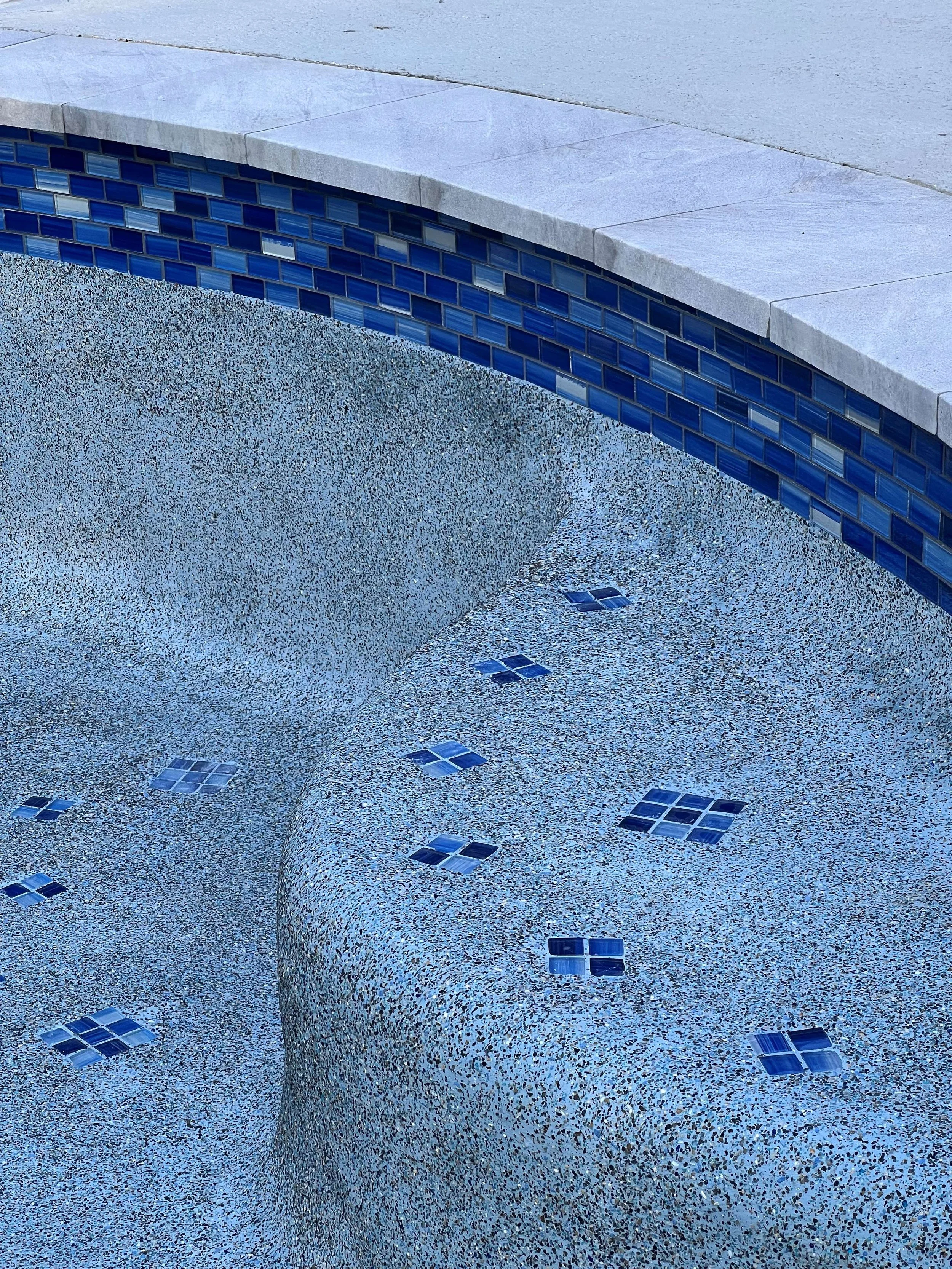 Close-up view of an empty swimming pool with a textured blue floor and small decorative blue tiles, with a stone-tiled edge.