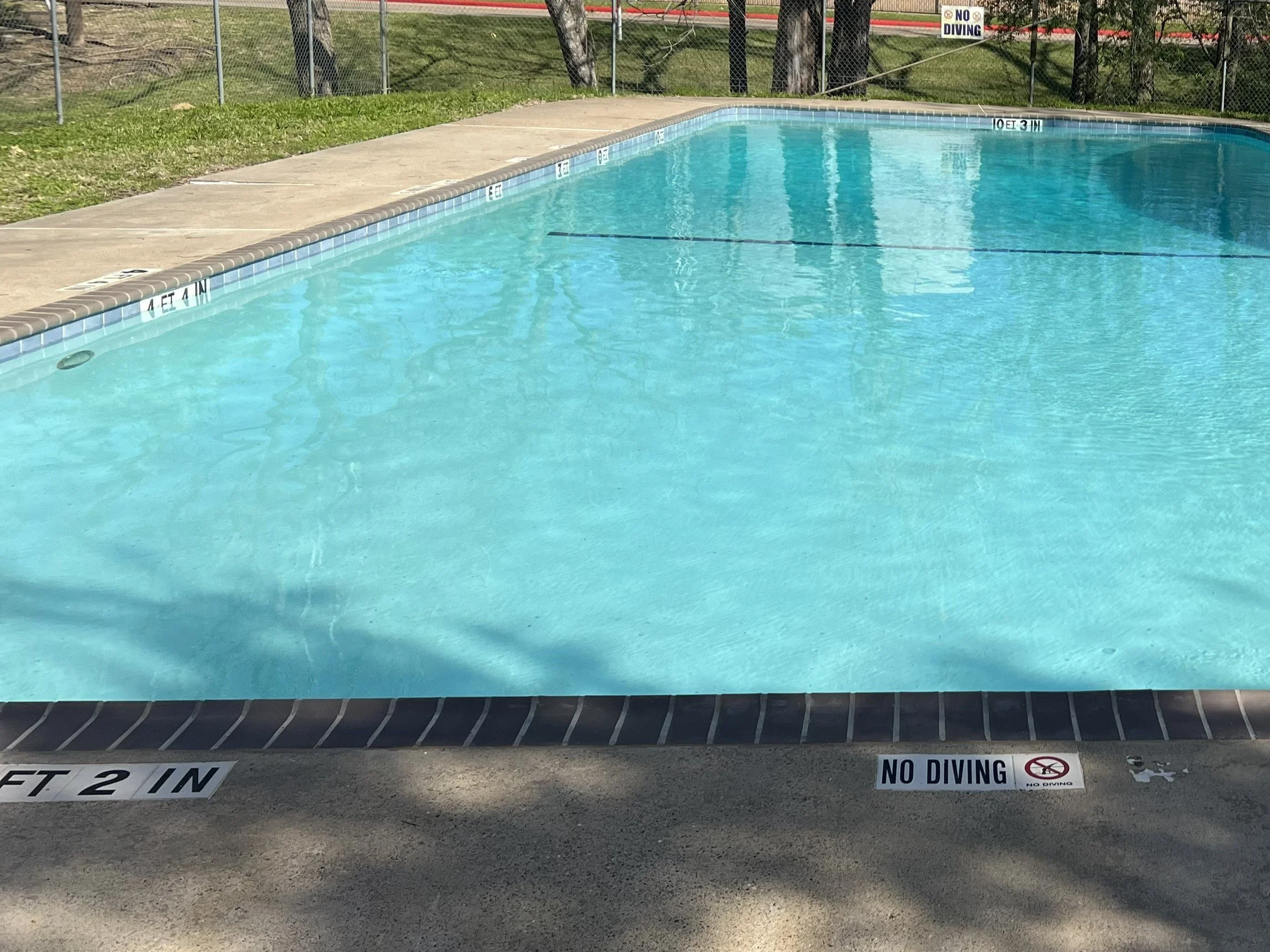 Empty swimming pool with clear blue water, surrounded by a concrete deck, and caution signs indicating no diving and no swimming.