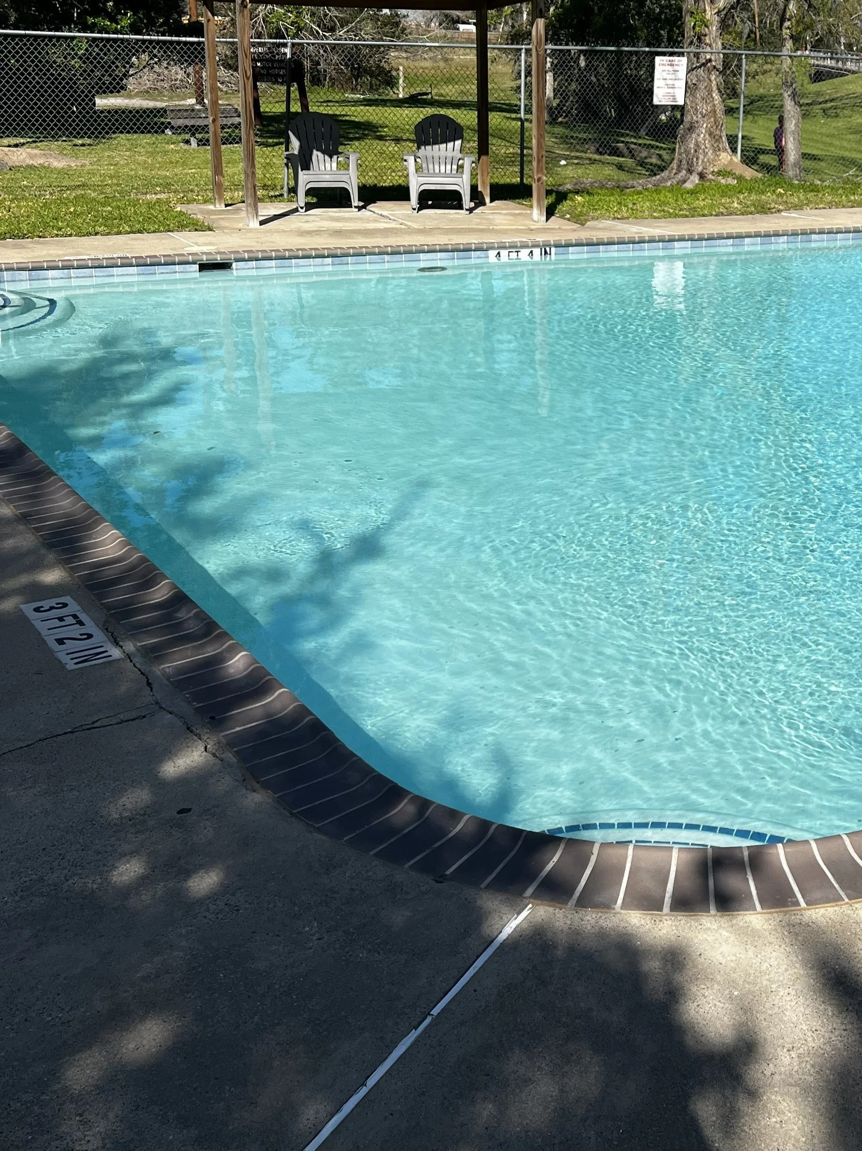 empty swimming pool with two white chairs on the pool deck, fenced area, green grass, trees, and a clear sunny day.