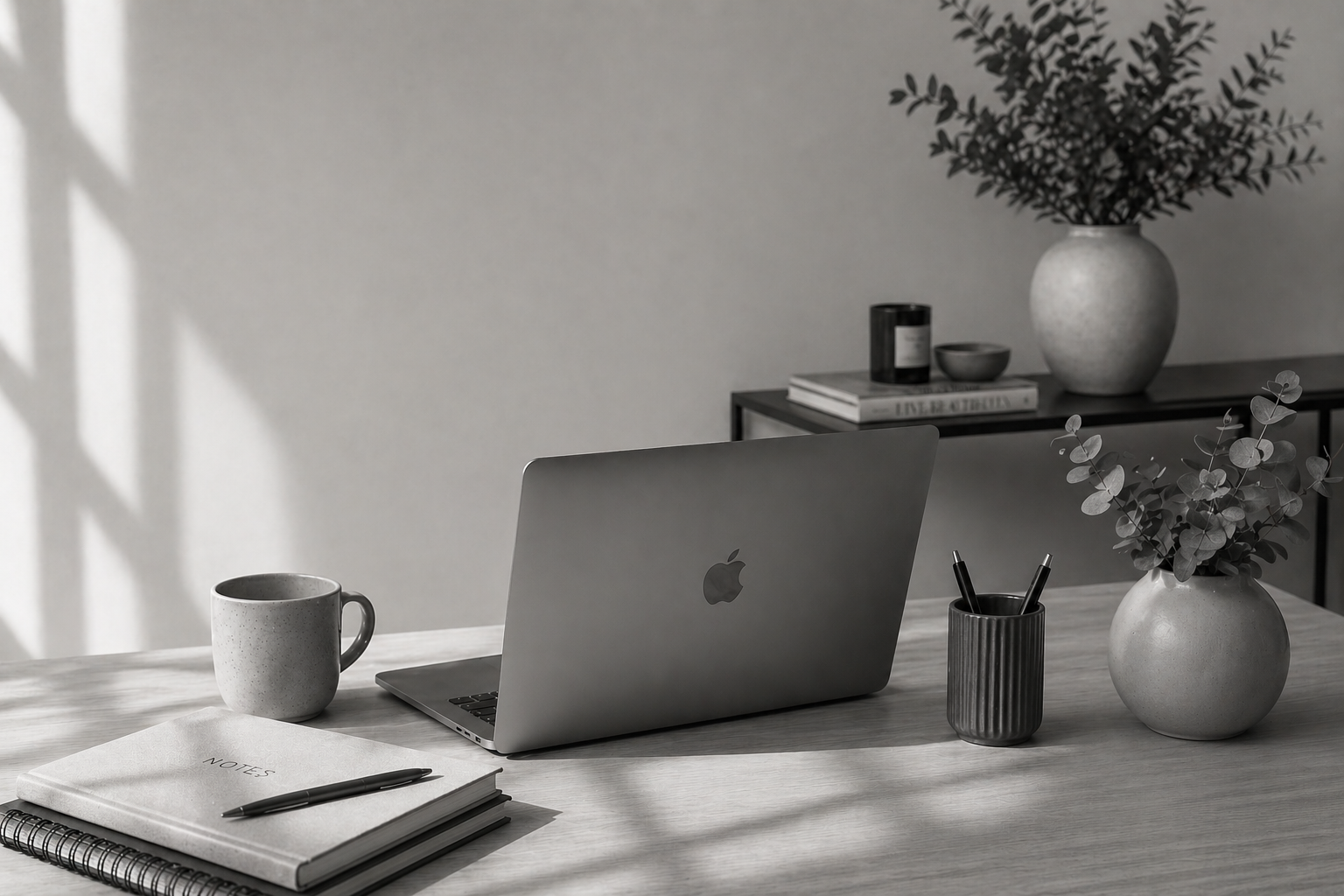 A workspace with a laptop, a notebook, a pen, a coffee mug, a vase with greenery, and a side table with books and a decorative item, in black and white.