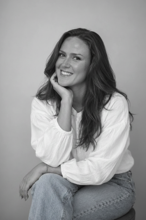 Black and white portrait of a woman with long wavy hair, smiling and resting her chin on her hand, sitting against a plain background.