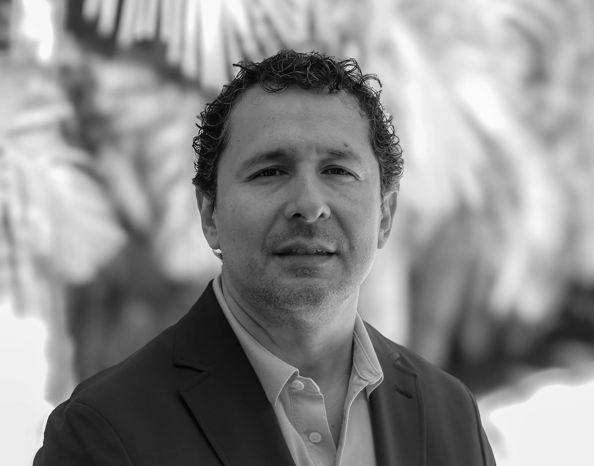 Black and white portrait of a man with curly hair wearing a suit and dress shirt, standing outdoors with a blurred background of trees and plants.