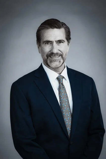 Professional portrait of a middle-aged man with dark hair, beard, and mustache, wearing a dark suit, white shirt, and patterned tie, facing forward against a neutral background.