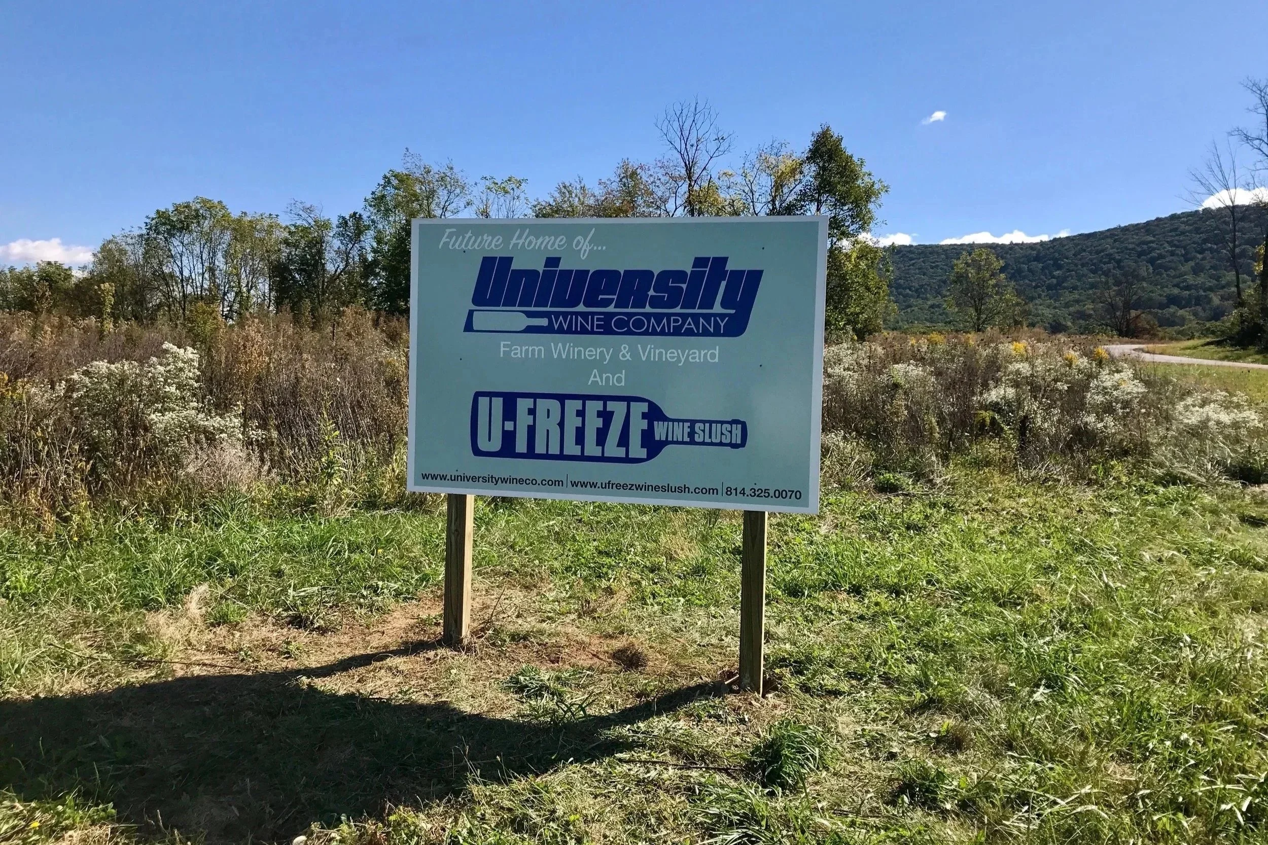 A sign on a grassy field promoting future home development of University Wine Company and U-Freeze Wine Slush, with trees and mountains in the background.