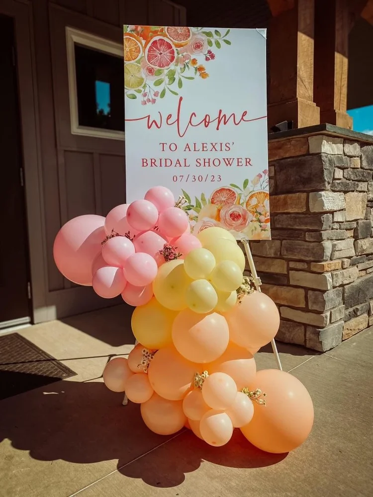 Decorative balloon arrangement with pink, peach, and yellow balloons in front of a welcome sign for Alexis' bridal shower on July 30, 2023, outside a building with a stone facade.
