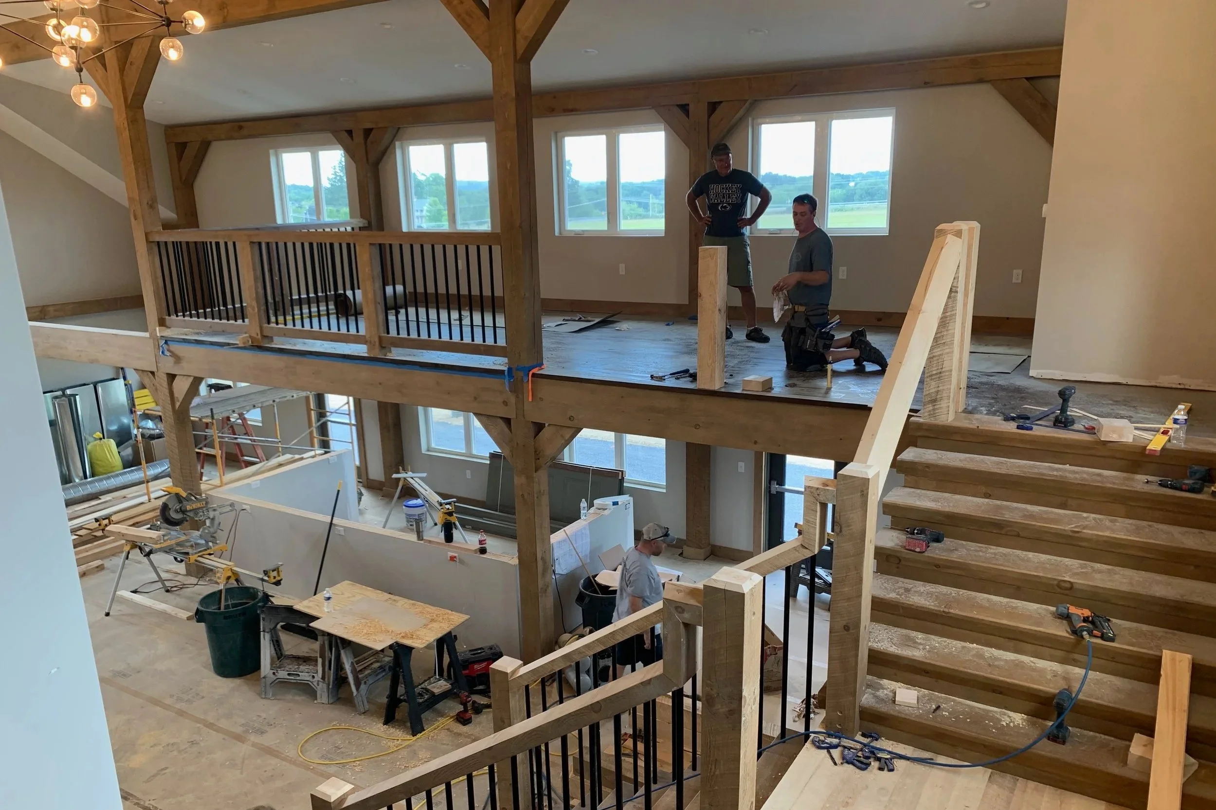 Two construction workers are installing hardwood flooring on the upper level of a house under renovation. The space has large windows, wooden beams, and a staircase leading to the lower level. Tools and construction materials are scattered around.