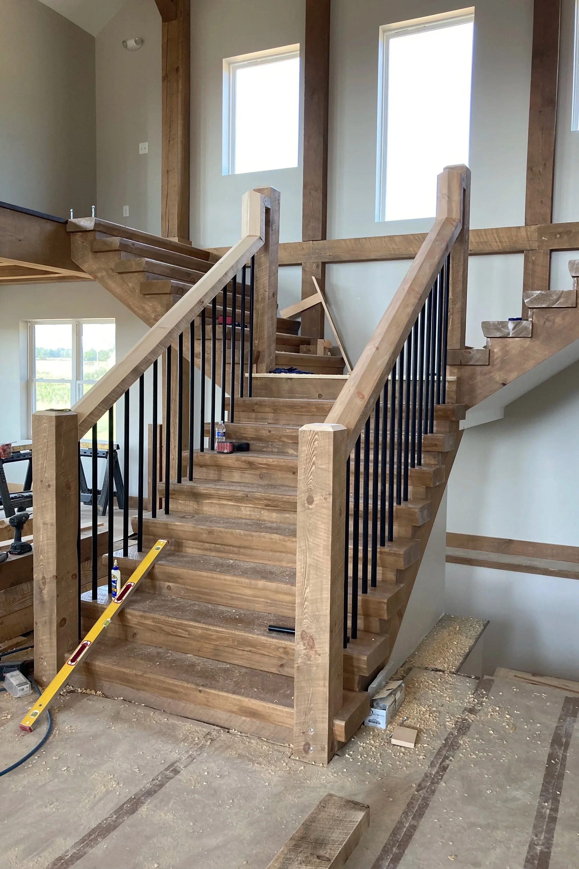 Interior of a house under construction, showing a wooden staircase with black metal balusters and unfinished stairs, surrounded by walls with large windows.