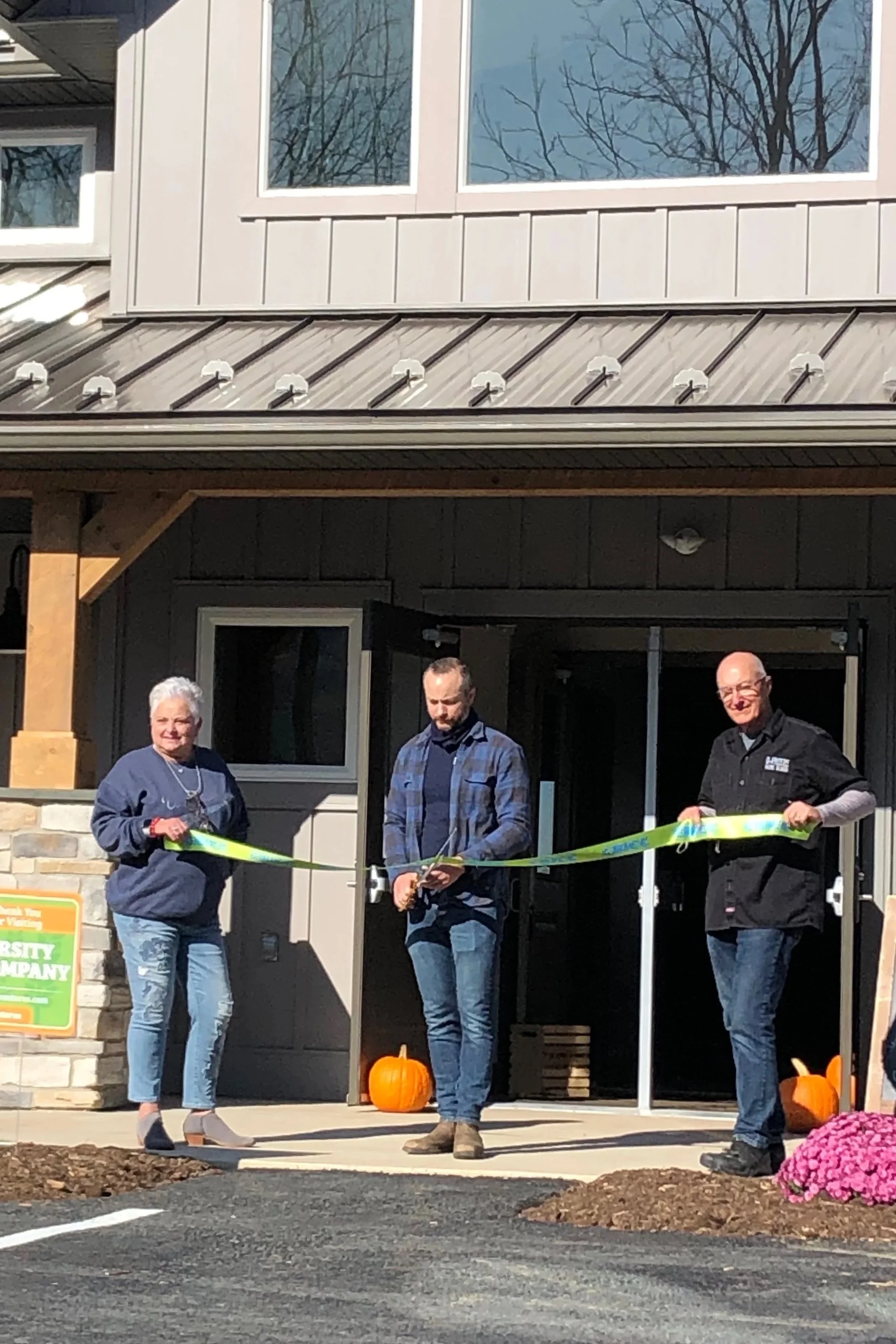Three people standing in front of a building with pumpkins, holding a ribbon for a ribbon-cutting ceremony.