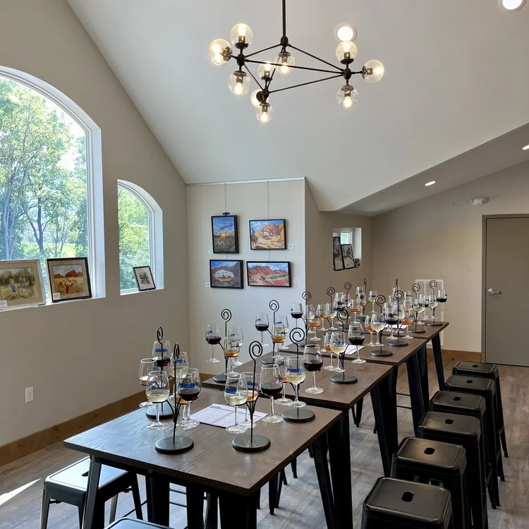 Room with high ceiling, modern chandelier, large windows, and a long table set with wine glasses and black hangers, likely for a wine tasting event.