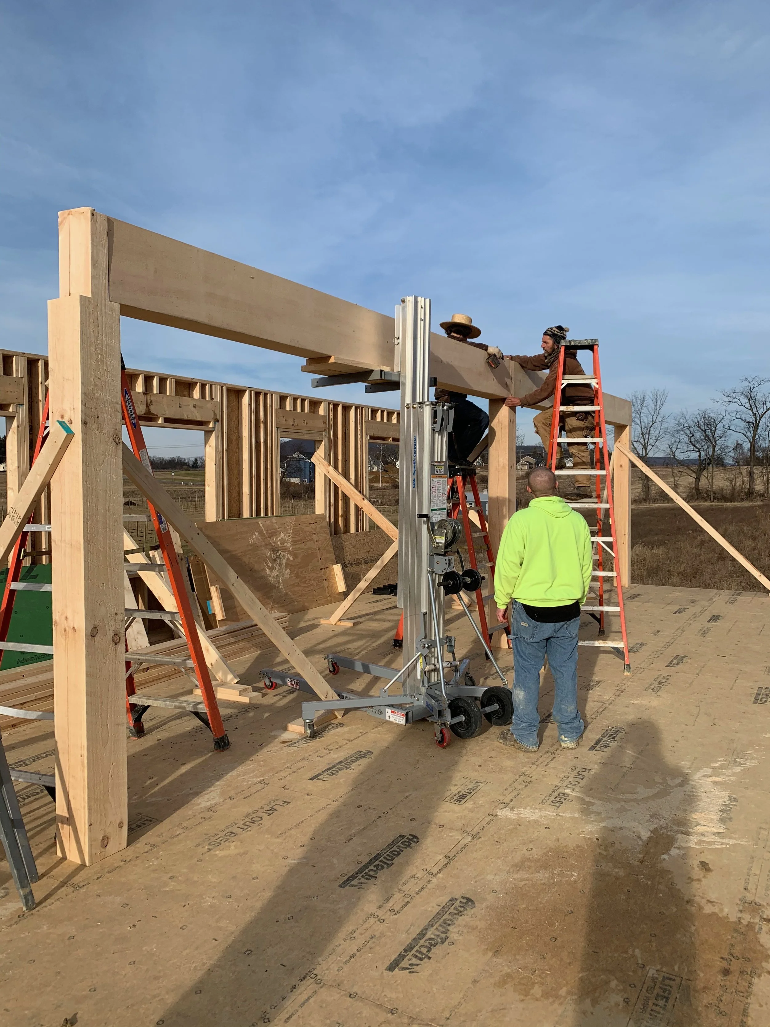 Construction workers building a house with wooden framing, some standing on ladders, with construction equipment on site, in an outdoor rural area under a partly cloudy sky.